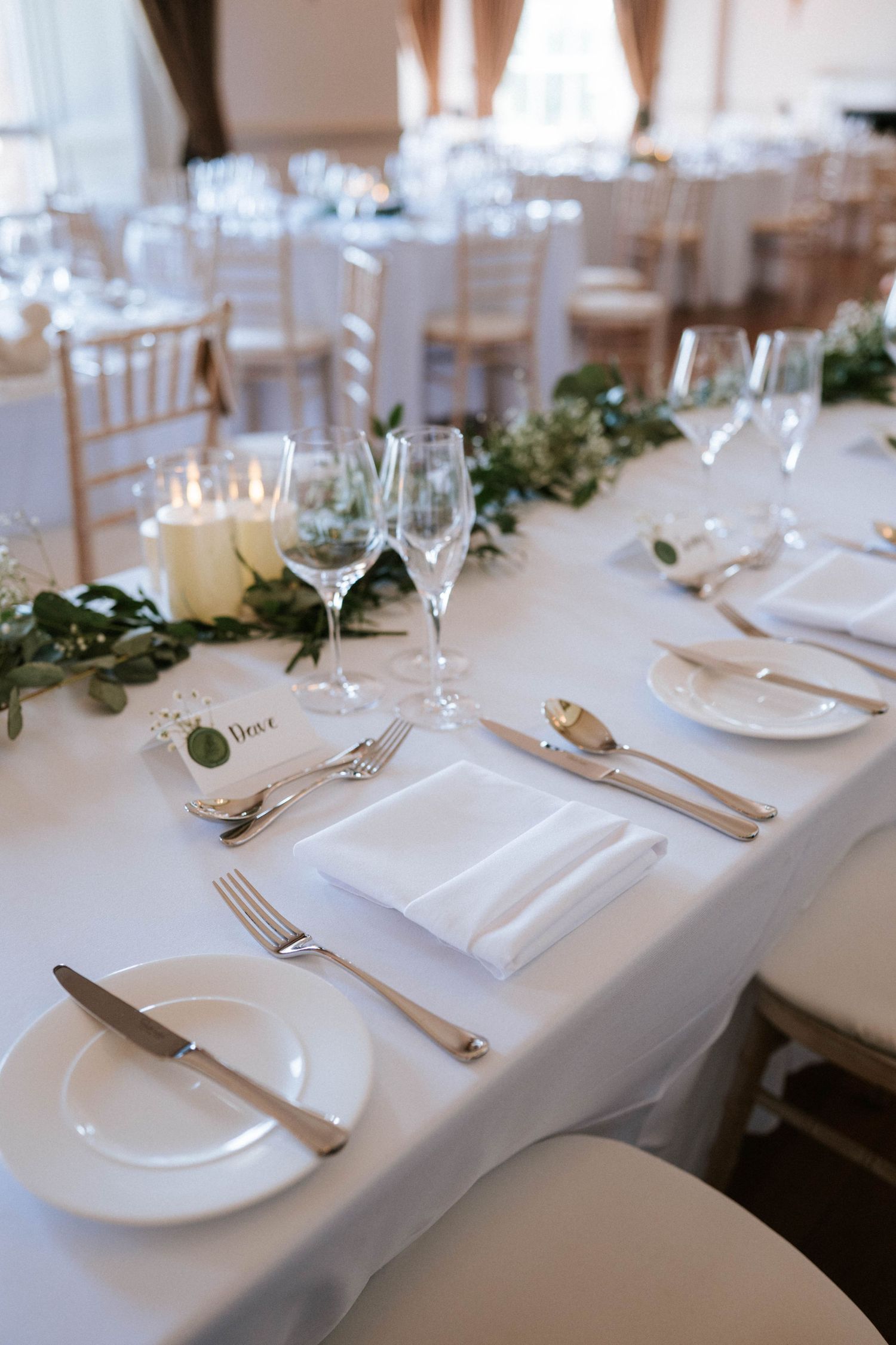 White place settings with silverware and wine glasses arranged on tables with greenery accents.