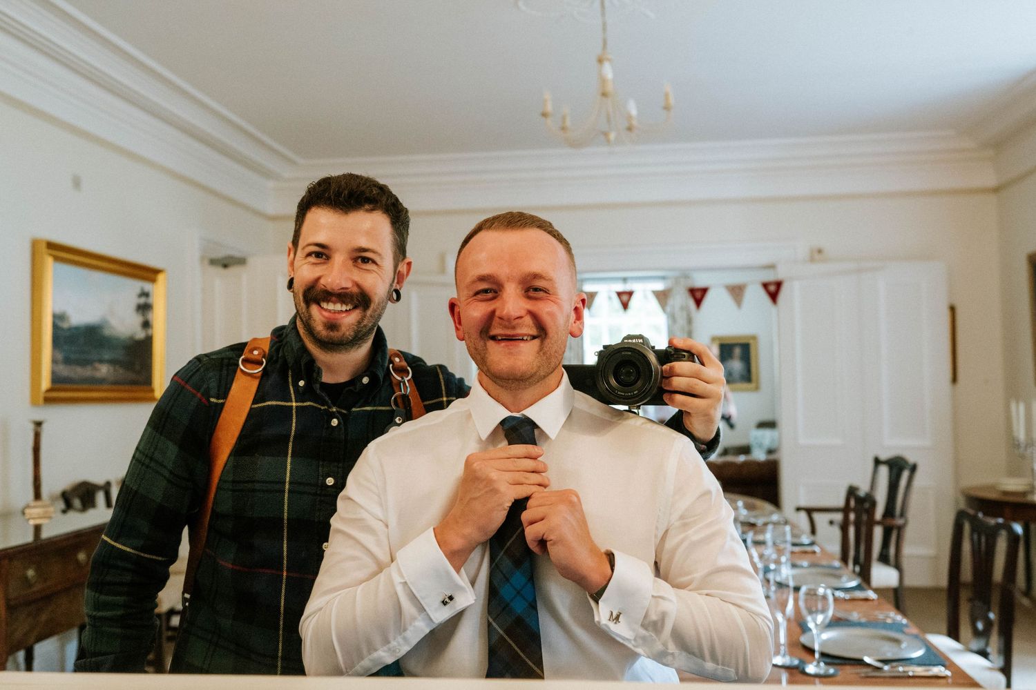 Two people smile together in a white interior room with glassware visible in the background.
