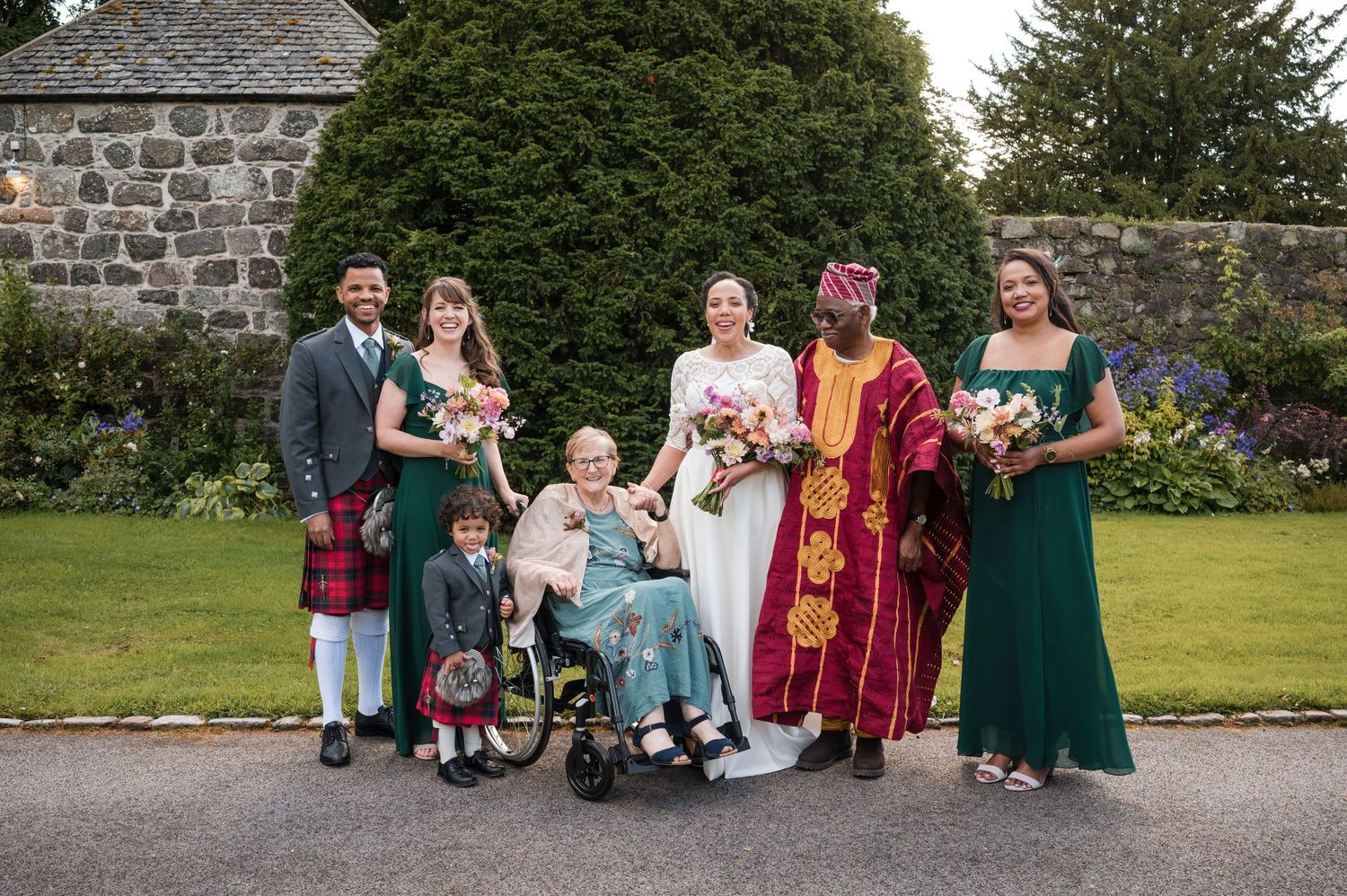 Wedding party posed for photos outside a stone building with greenery.