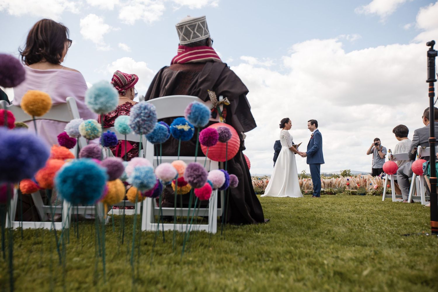Outdoor ceremony setup with colorful pom poms and white chairs on a hilltop.