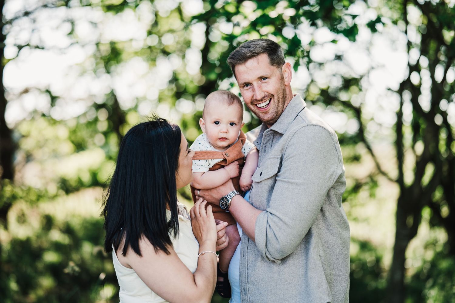A couple shares joyful laughter together in a sunny outdoor setting with lush greenery in the background.