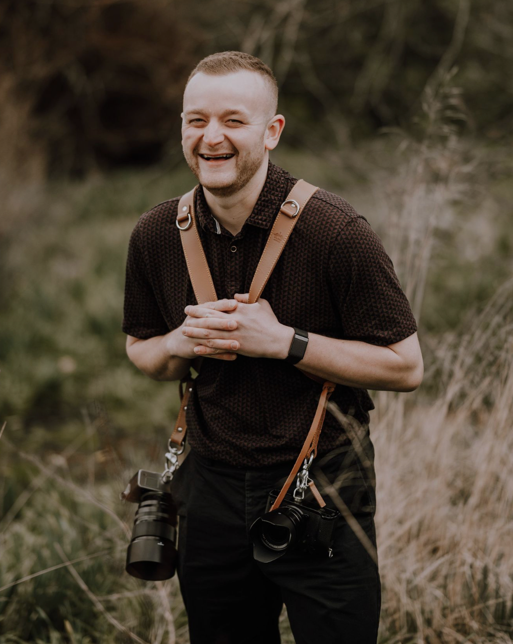 Professional photographer in dark attire smiles while holding camera equipment in outdoor natural setting.