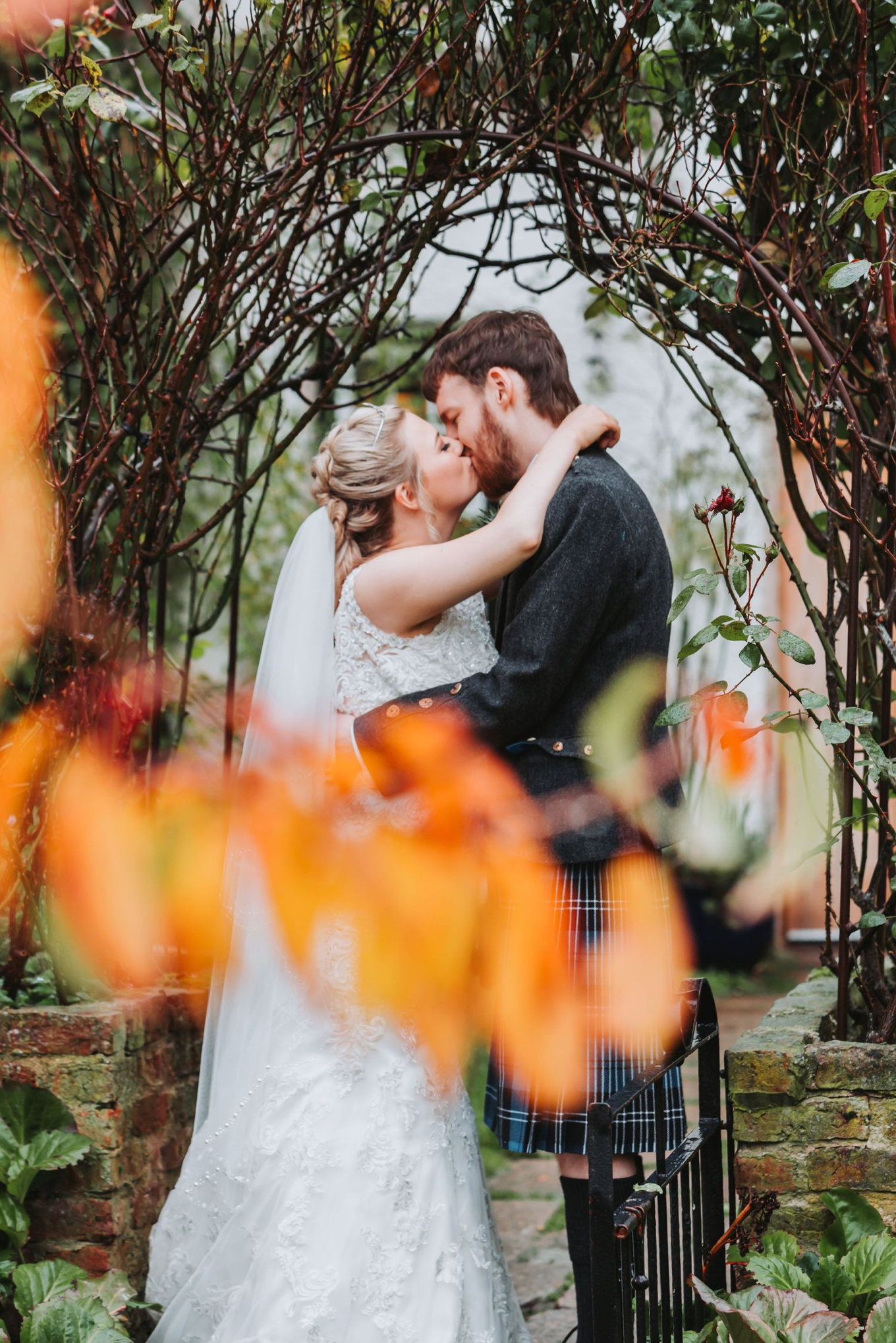 A couple shares a romantic kiss under a floral arch with orange flowers blooming in the foreground.