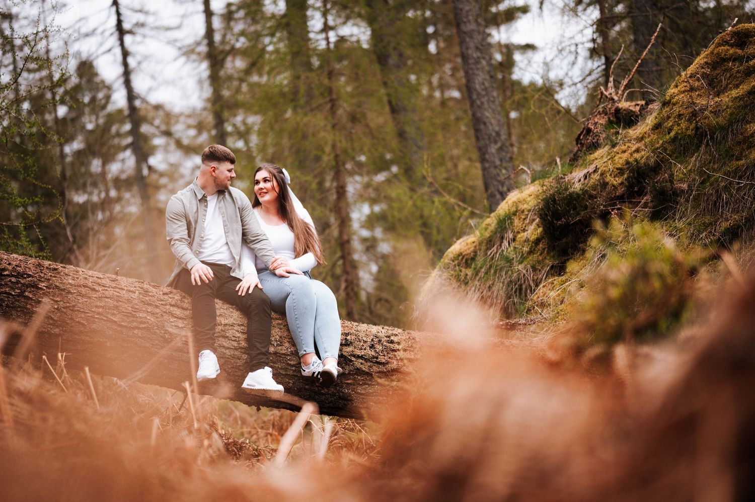 A couple shares a romantic moment sitting together on rocks in a wooded forest setting during sunset.