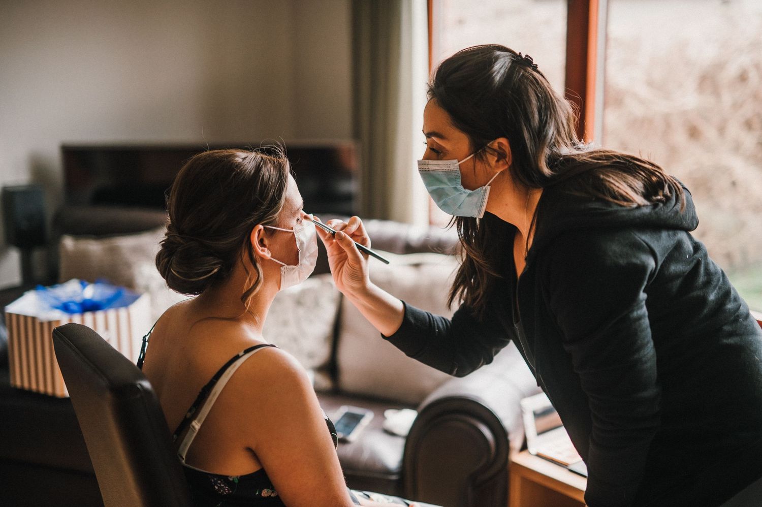 Makeup artist applying cosmetics during bridal preparation while wearing protective face masks.