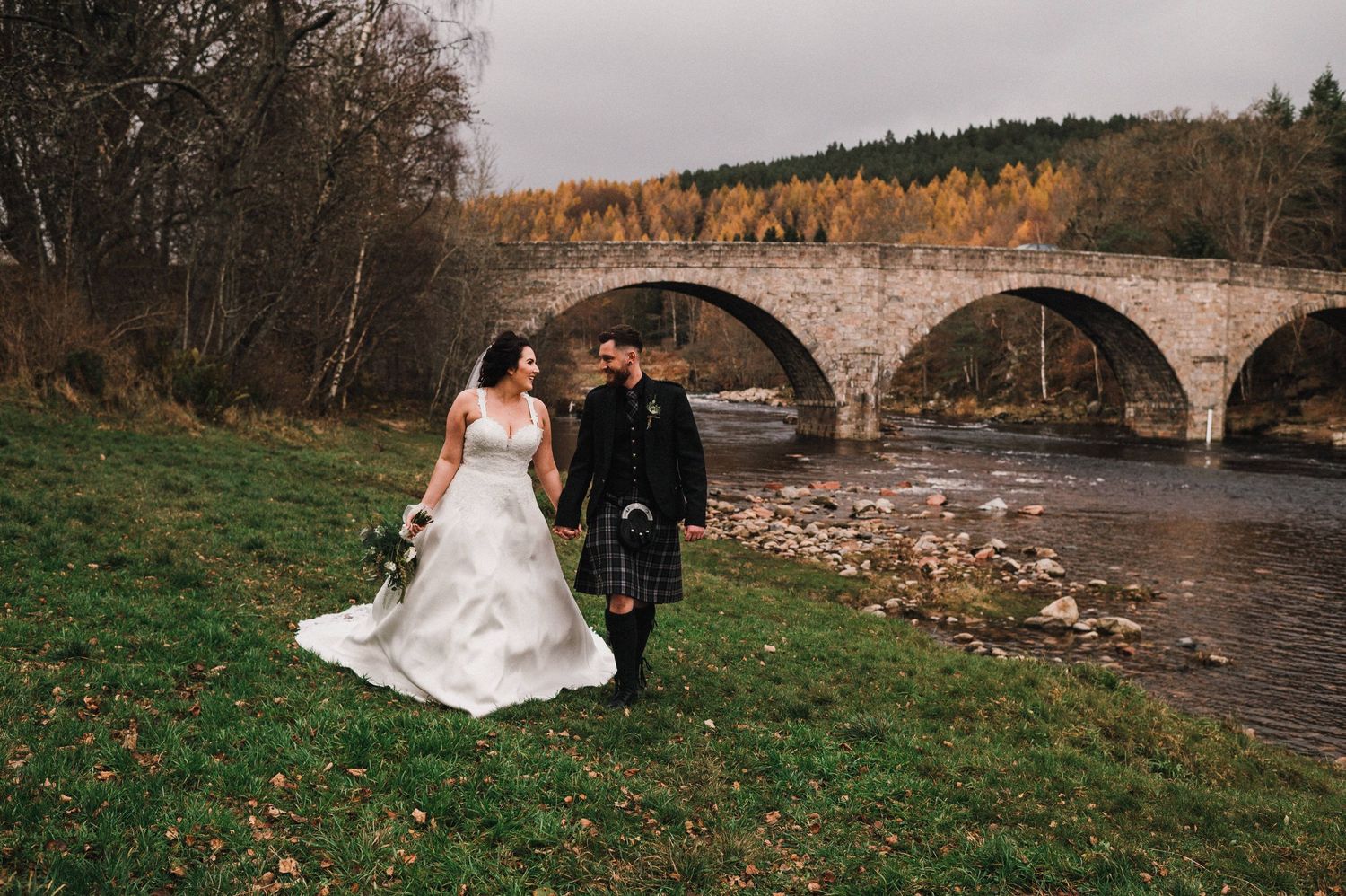 Newlyweds in Scottish wedding attire pose by historic stone bridge in autumn setting.