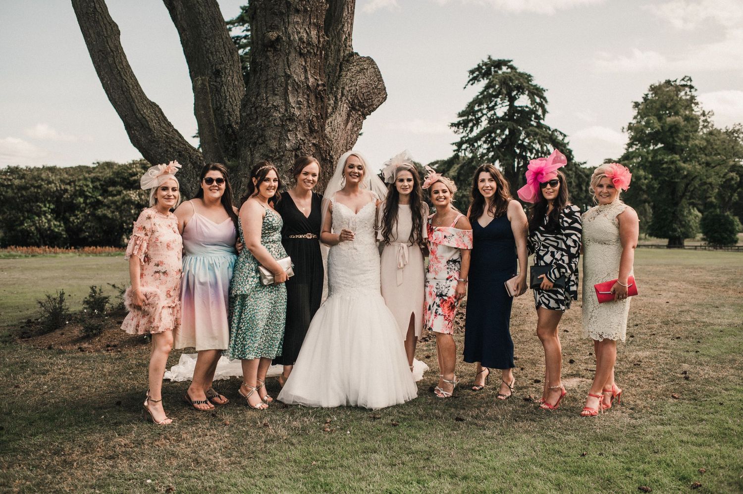 A wedding party of women in colorful dresses poses on a grassy lawn with trees in the background.