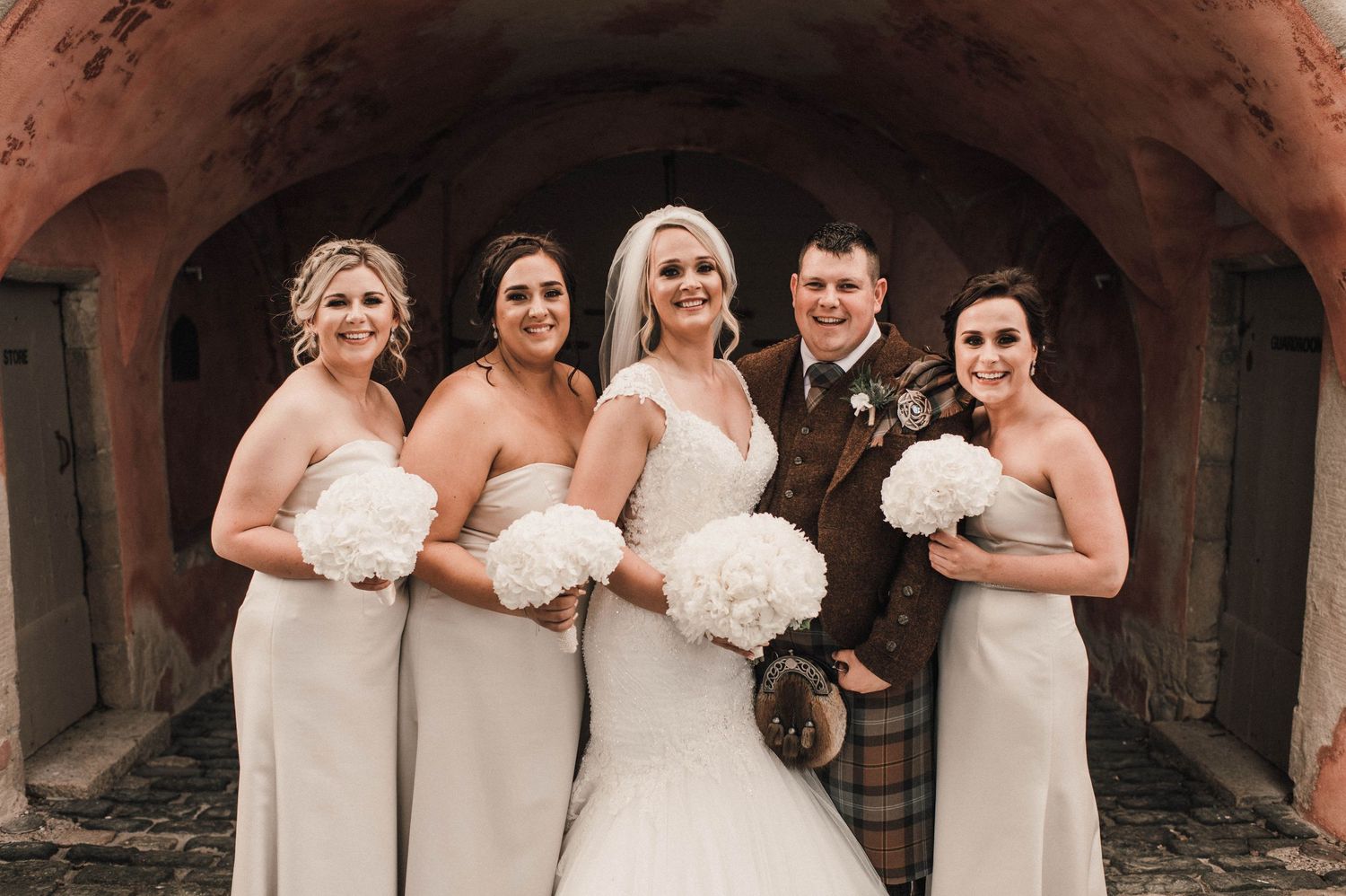 Bridal party poses under rustic stone archway wearing neutral colored gowns and holding white bouquets.
