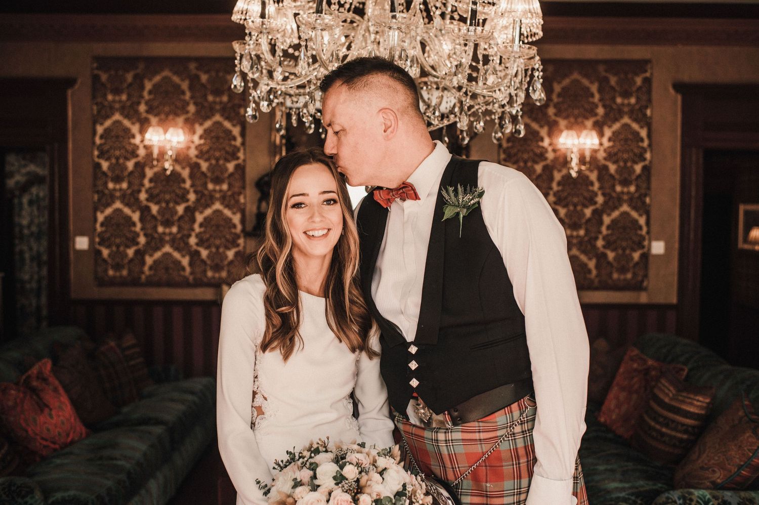 Intimate moment between newlyweds under ornate chandelier in traditional Scottish wedding attire.