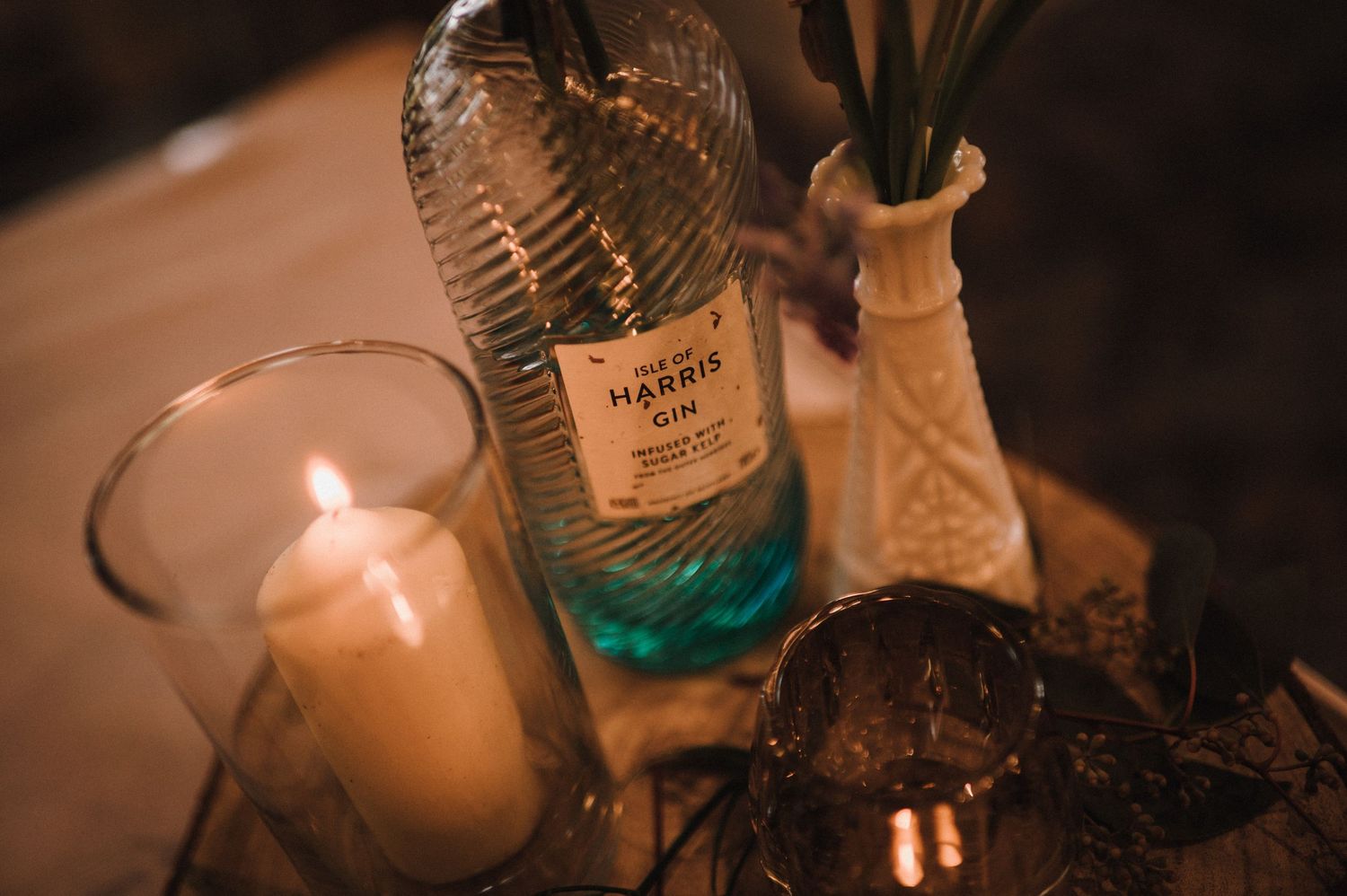 Glass bottle and candle centerpiece with delicate floral arrangement on wooden surface.