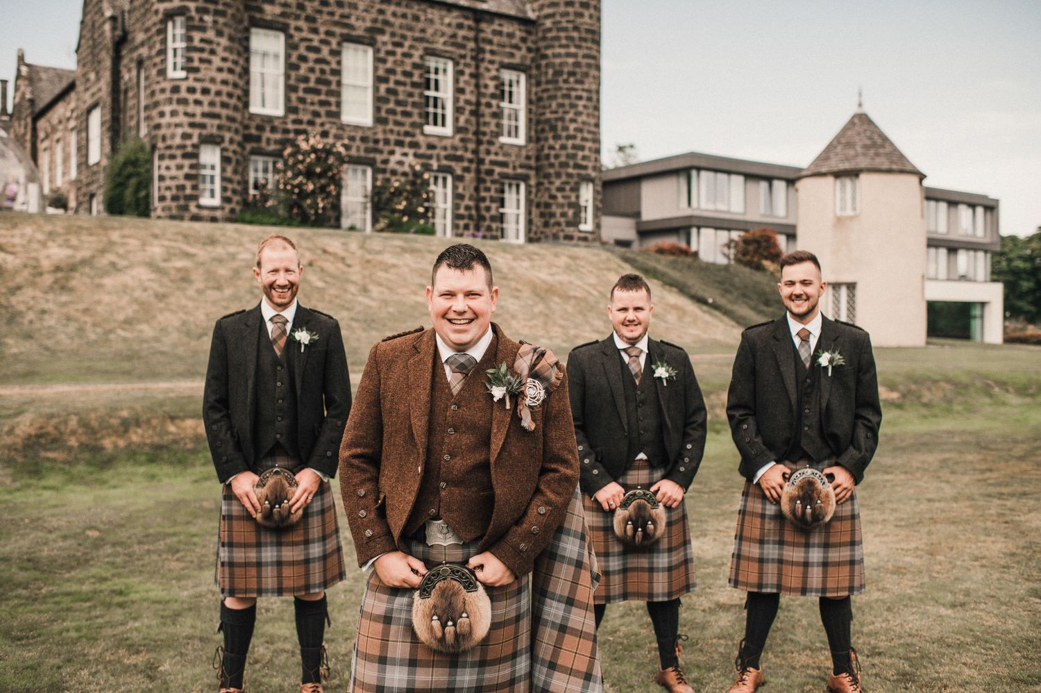 Wedding party in kilts and formal attire pose in front of stone building.