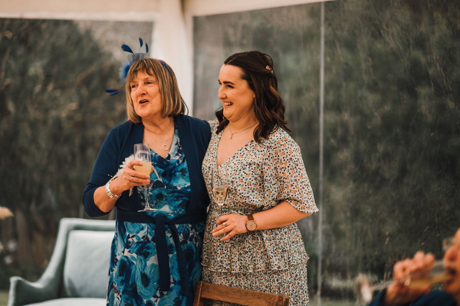 Two guests in festive dresses share drinks and laughter at an outdoor celebration.