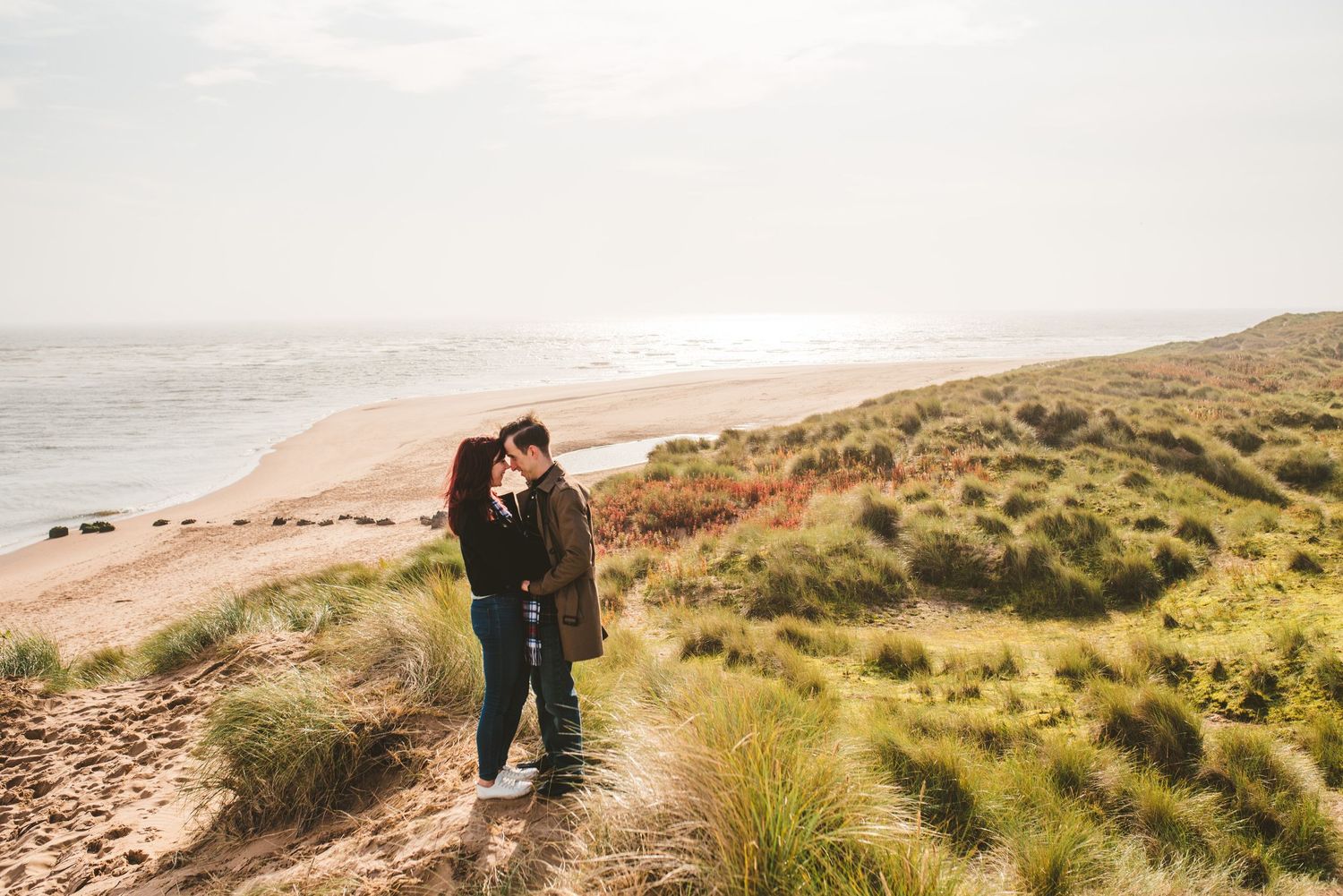 Couple embracing on coastal clifftop path overlooking scenic beach at sunset.