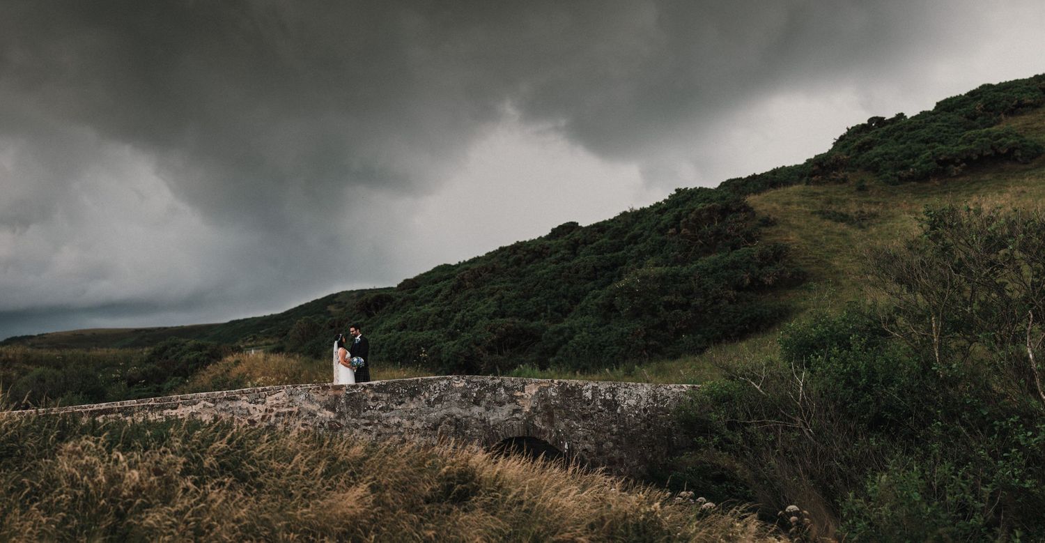 A dramatic wide-angle wedding photo on a stone path with dark storm clouds overhead.