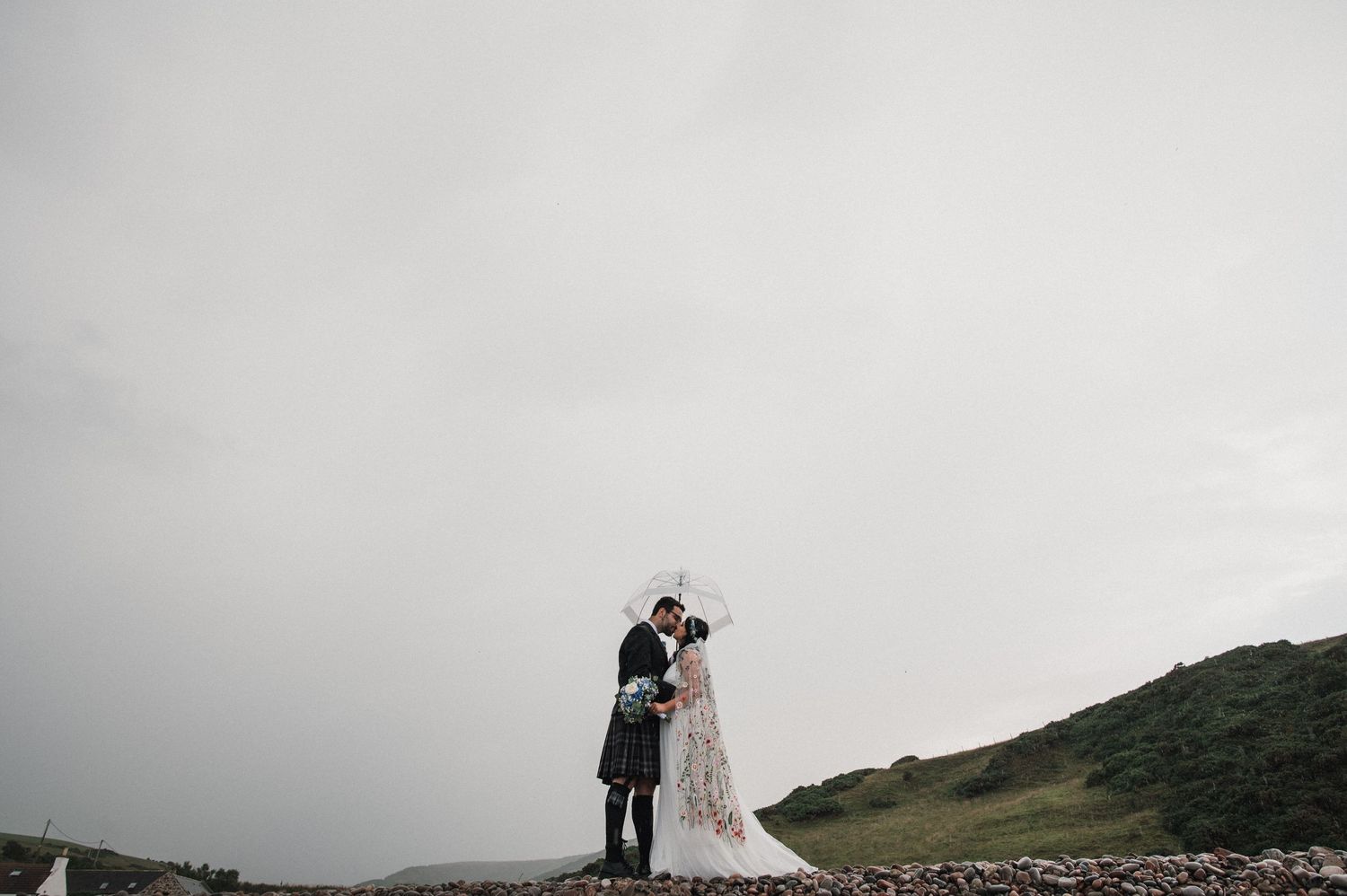 A distant view of newlyweds embracing on a hilltop against a moody gray sky.