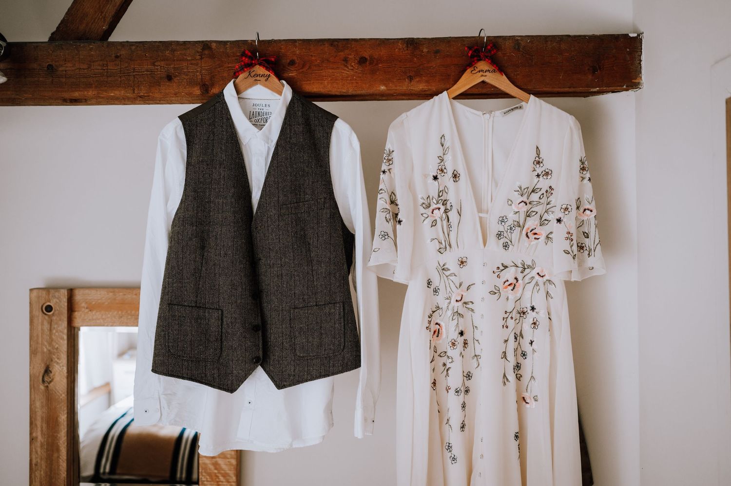 A grey tweed vest and white floral wedding dress hanging on wooden beams.