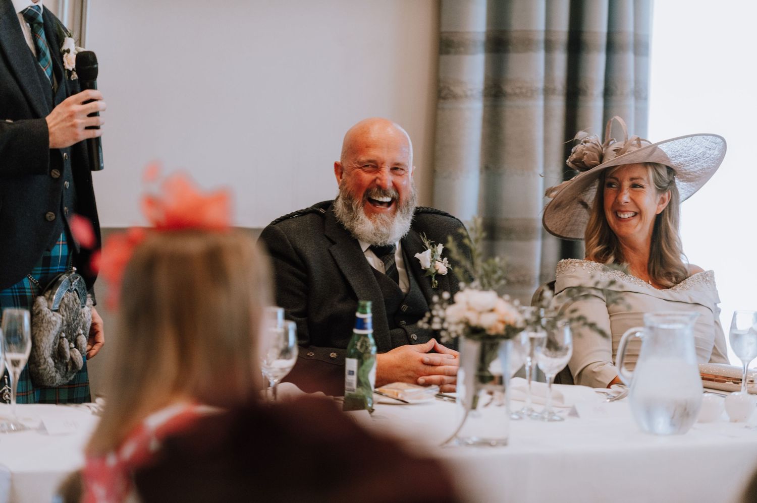 Wedding guests at a formal reception share laughter during speeches.