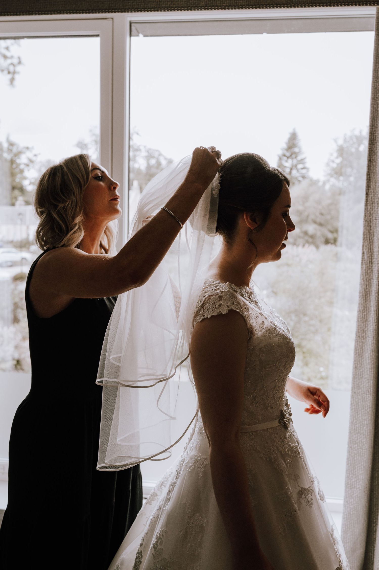 A bridal veil being carefully adjusted in front of large windows with natural lighting.