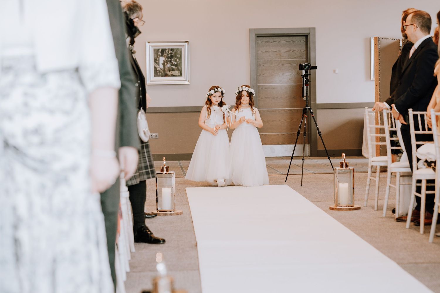 A bridesmaid in a long coral dress walks down a white aisle runner carrying a floral bouquet.