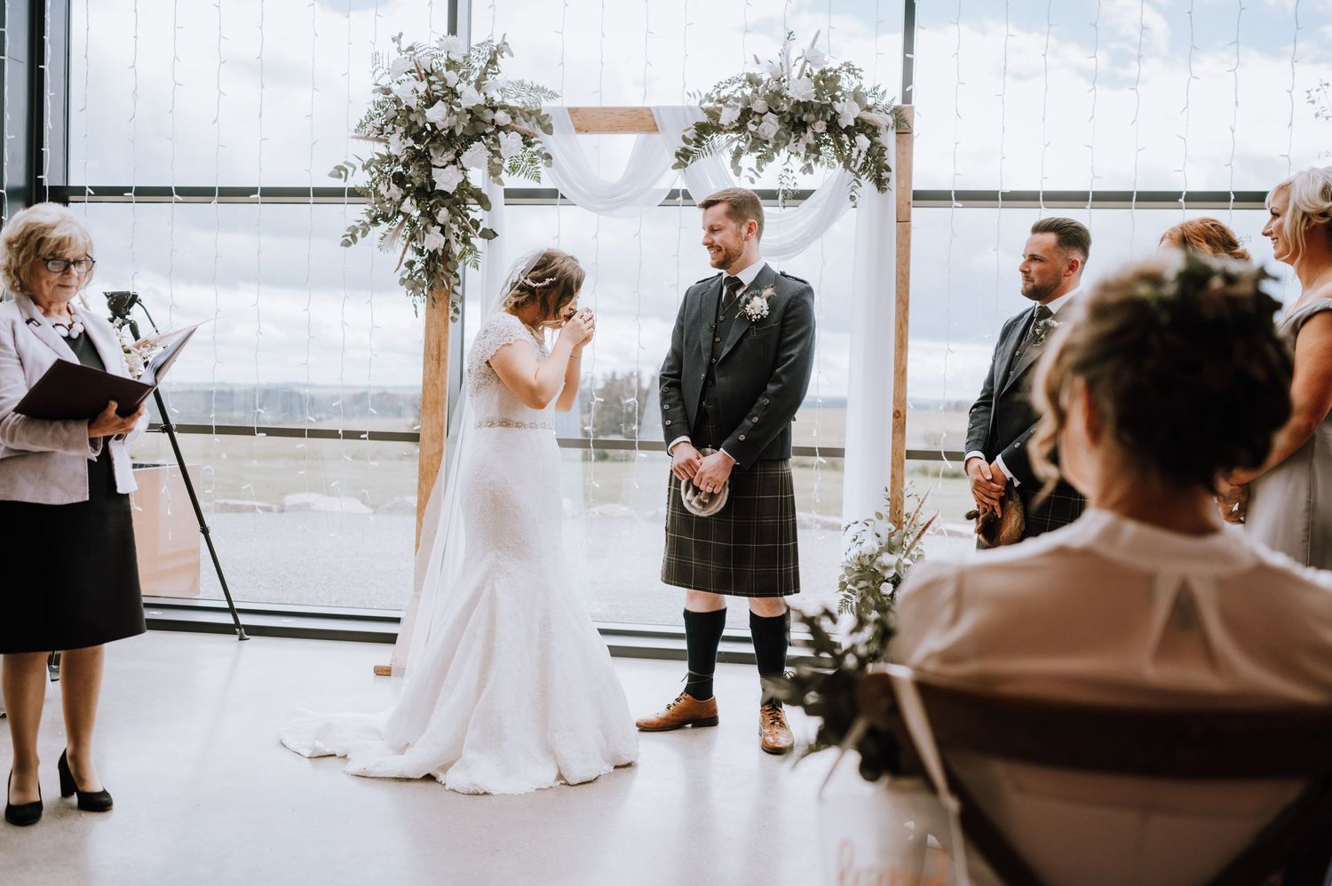 A Scottish wedding ceremony in a modern venue with large windows and floral arch decorations.
