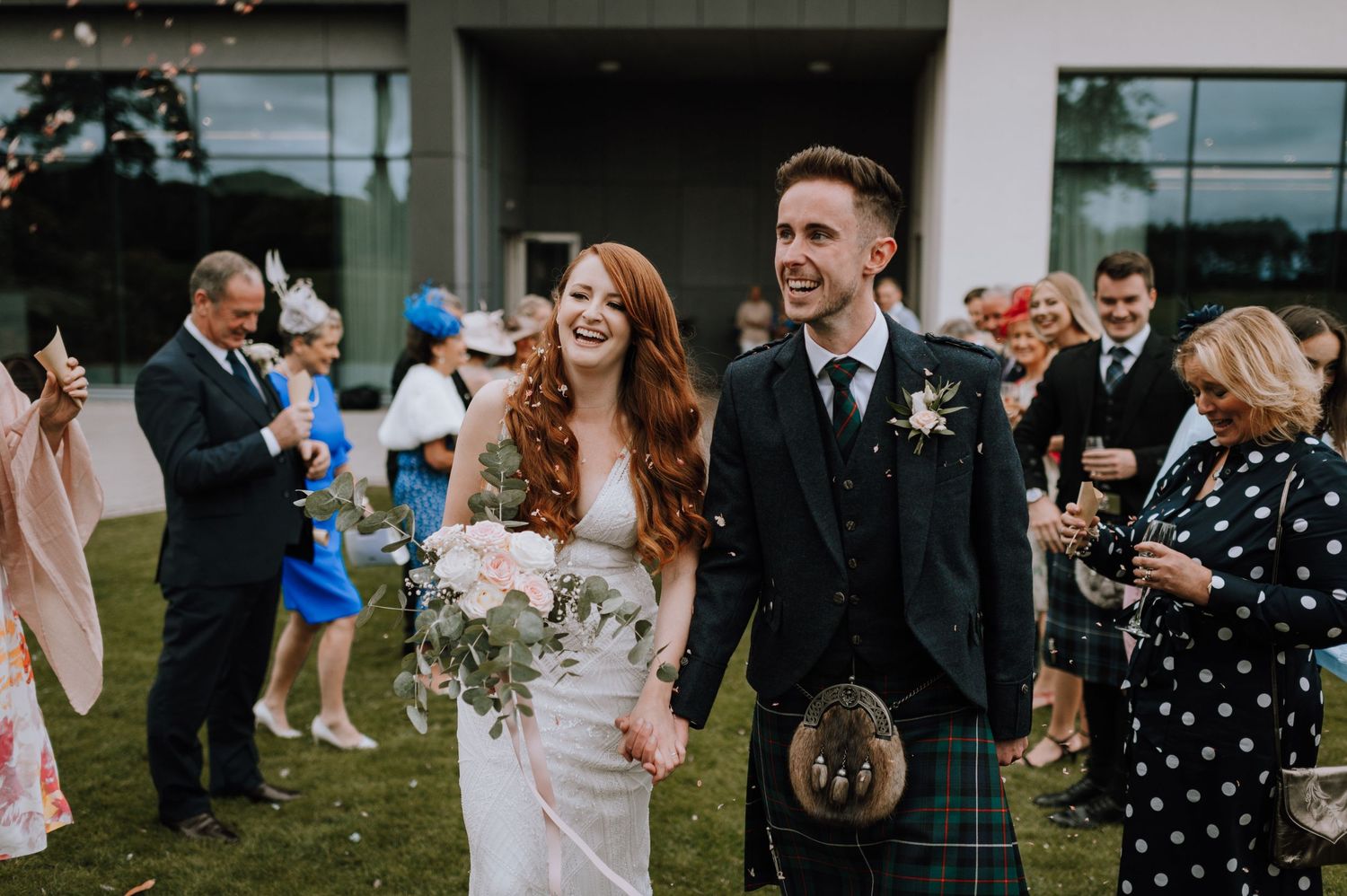 Guests celebrate during a wedding with the couple in Scottish attire walking through confetti.