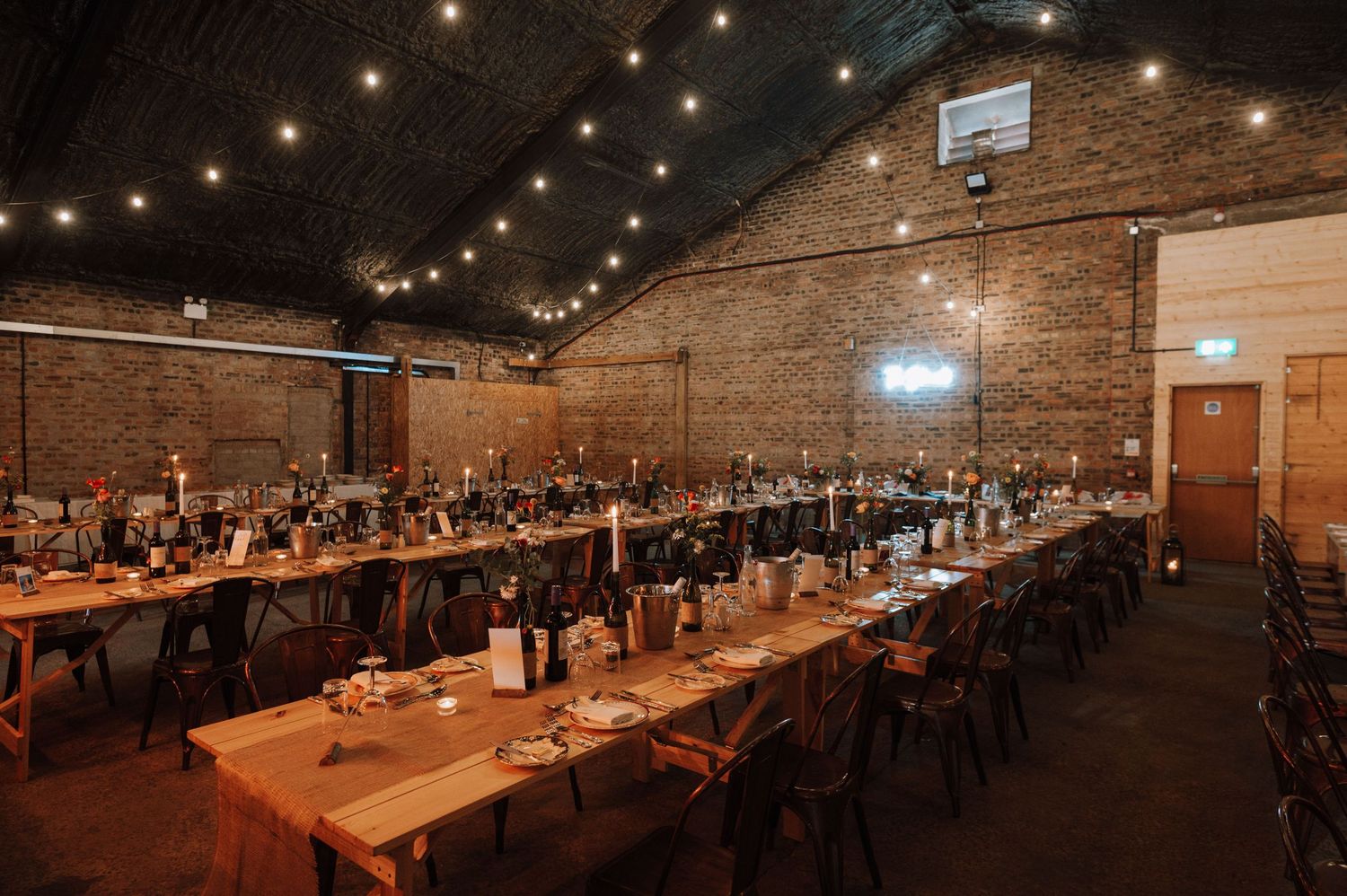 Large rustic wedding venue with exposed brick walls, string lights and long wooden dining tables.