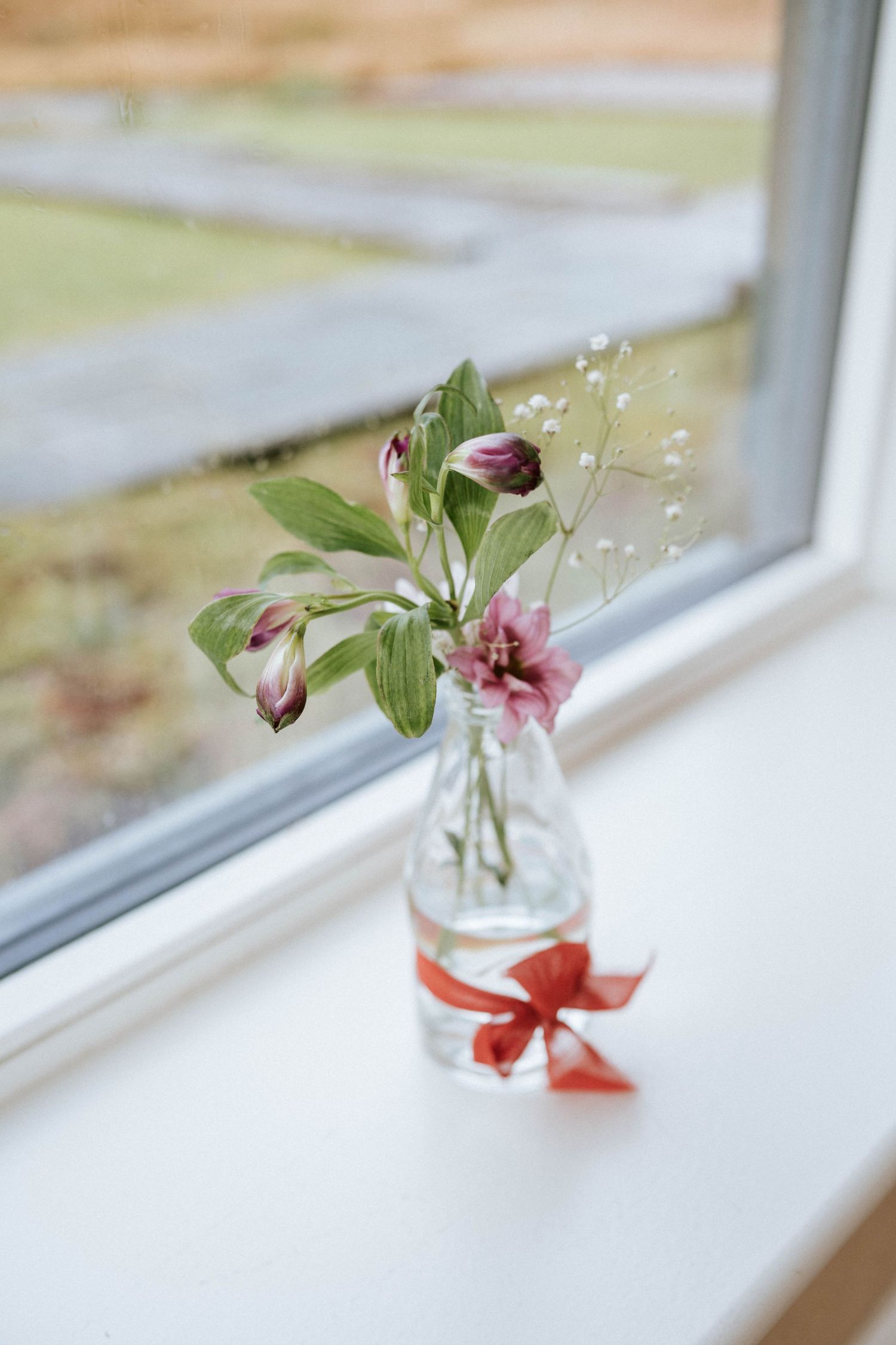 Pink flowers in a clear glass vase with red ribbon sitting on a windowsill.