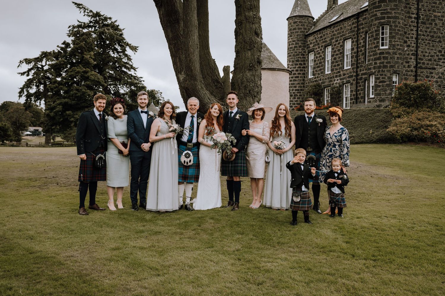 Formal group photo of wedding party standing together on castle grounds wearing traditional Scottish attire.