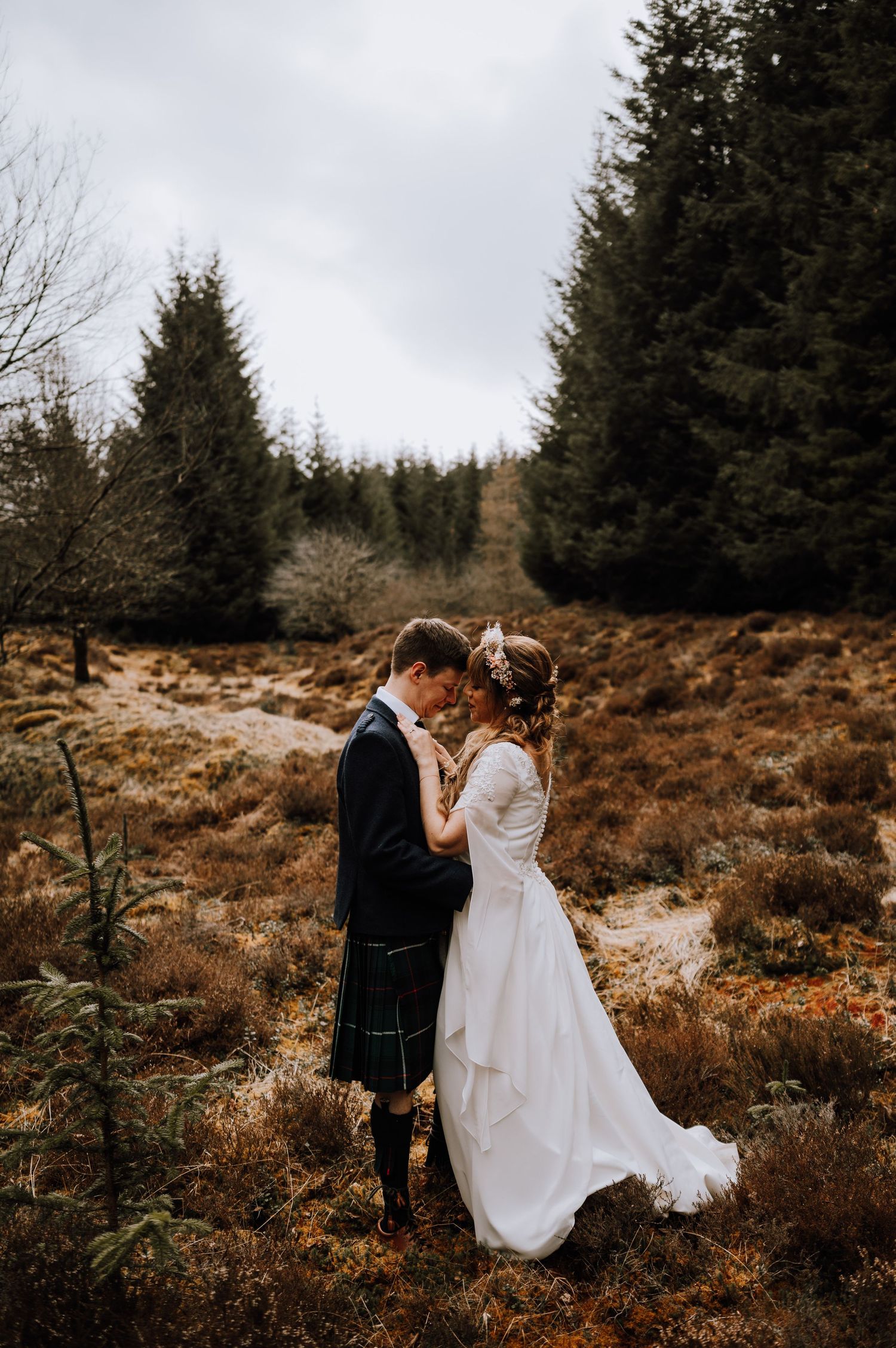 Intimate wedding portrait of couple embracing in scenic Scottish Highlands among evergreen trees.