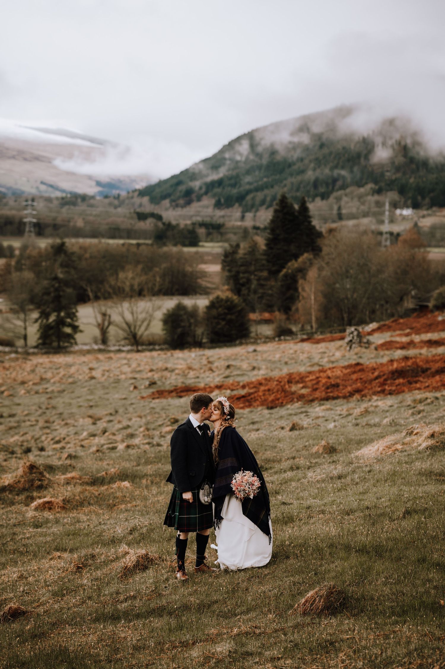Couple shares intimate moment on misty Scottish hillside in traditional wedding attire with mountains in background.