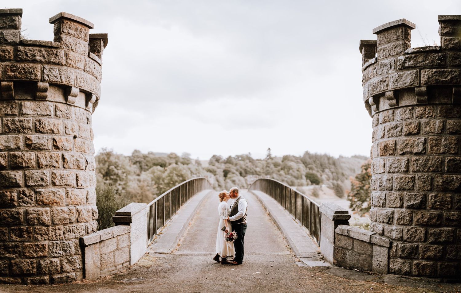 A couple poses between stone castle turrets on a bridge during their wedding photoshoot.