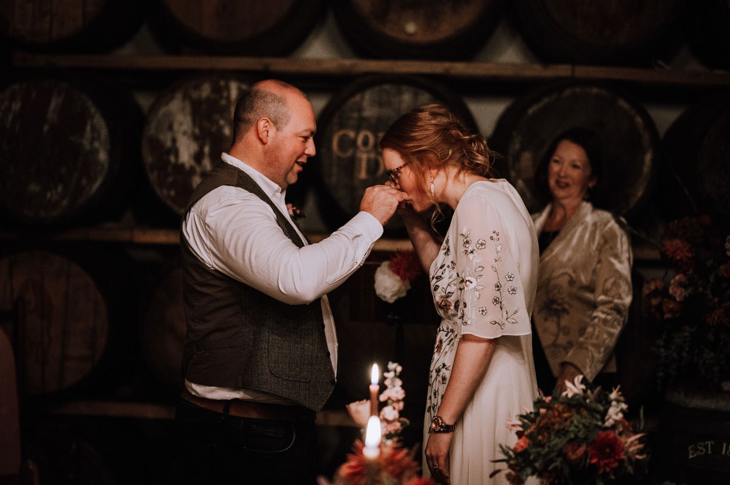 Emotional moment between couple sharing a tender gesture in a rustic wine cellar with wooden barrels in background.
