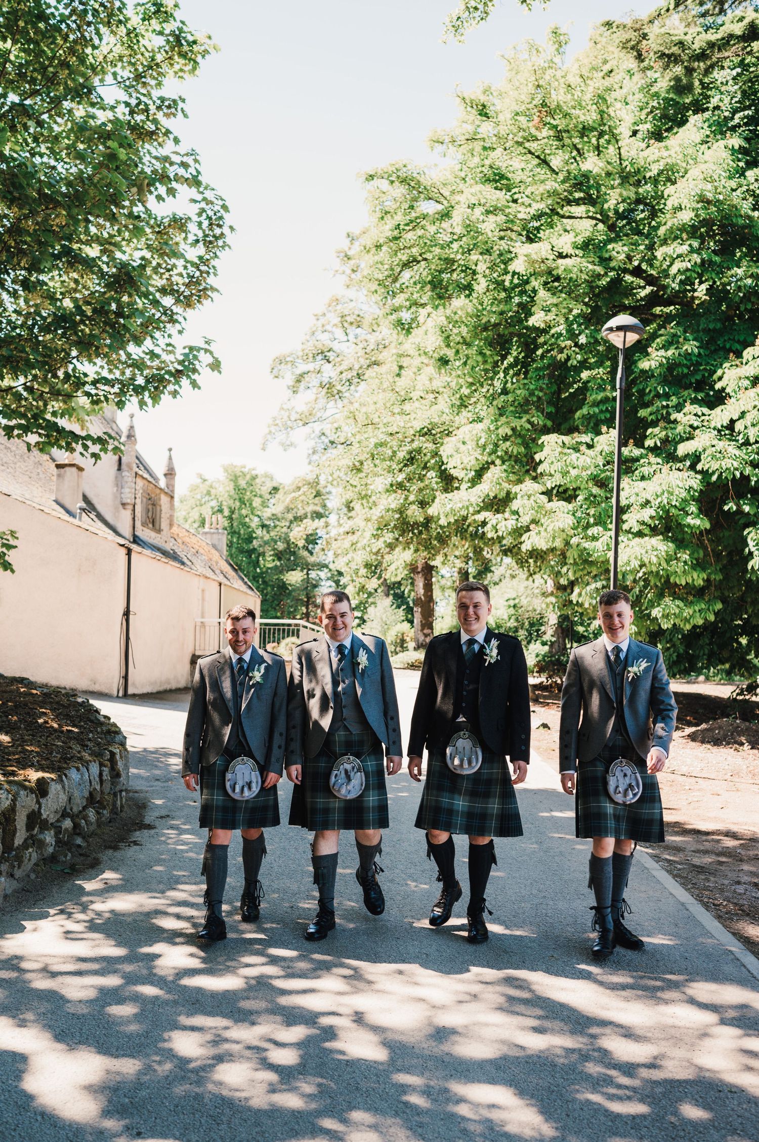 Four people in matching kilts and formal attire stand together on a tree-lined path.