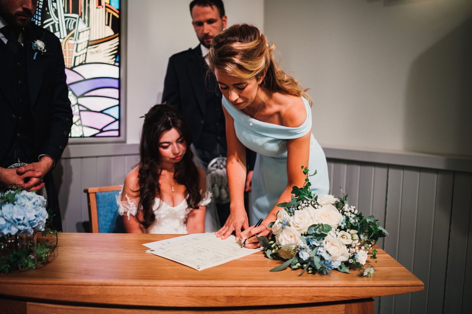 Wedding document signing with light blue bridesmaid dress and white floral arrangements on wooden table.