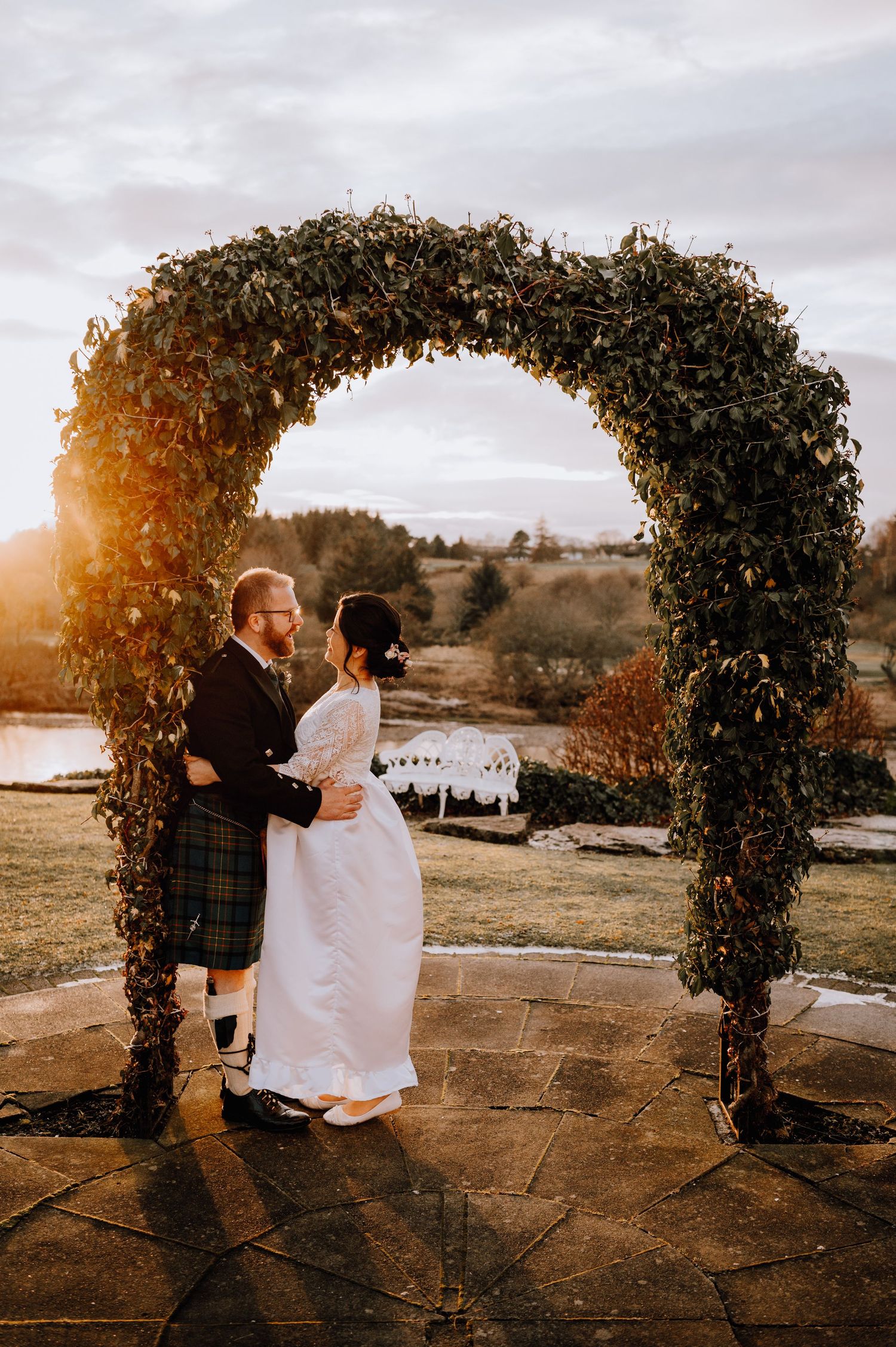 Newlyweds share a romantic moment under a lush floral arch during golden hour at their outdoor ceremony.