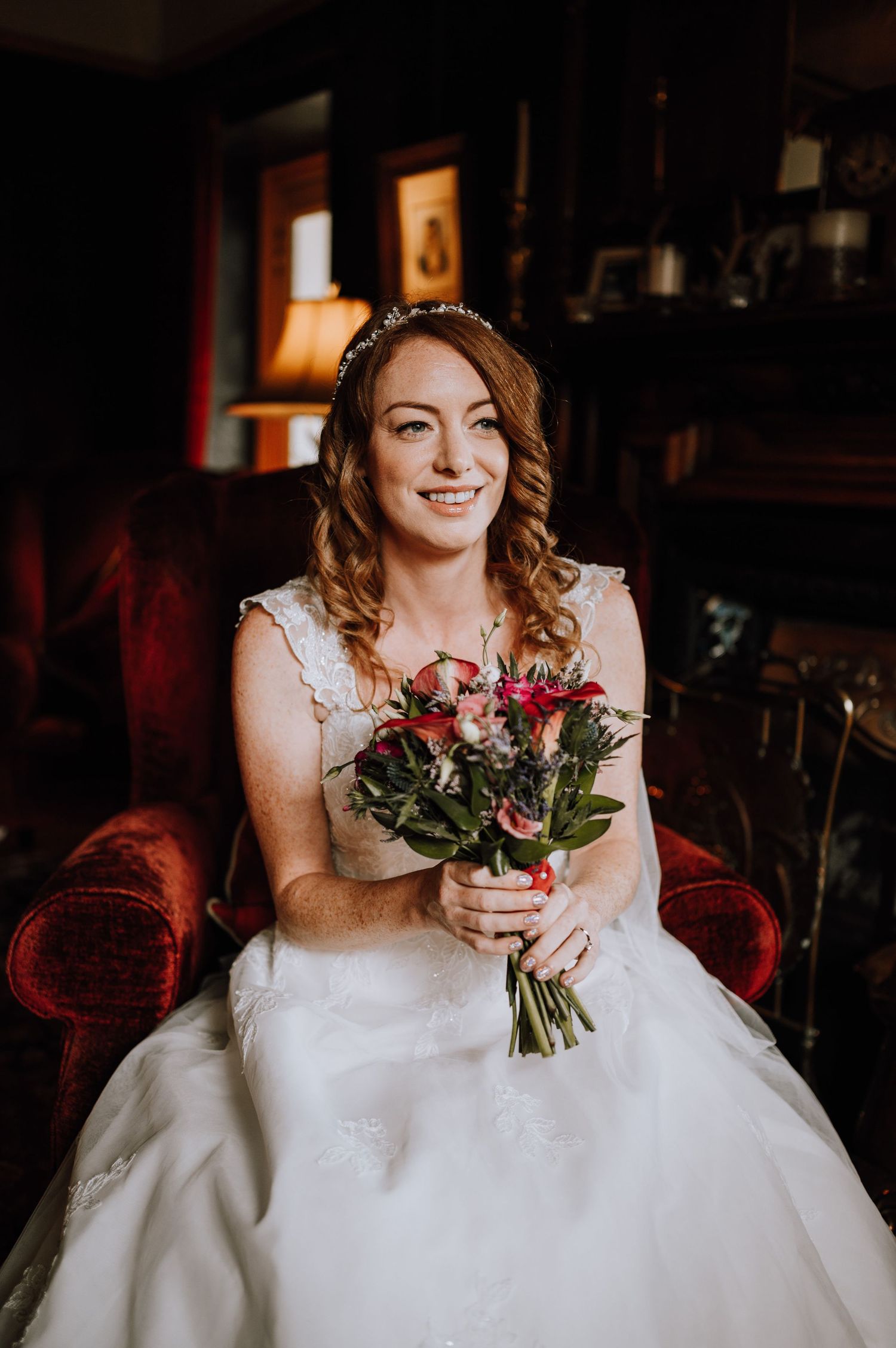 Bride in white dress holding pink bouquet while sitting on red velvet chair.