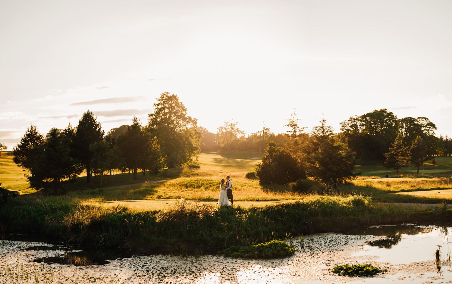 A scenic wedding photograph of a couple silhouetted against a golden sunset near a pond.