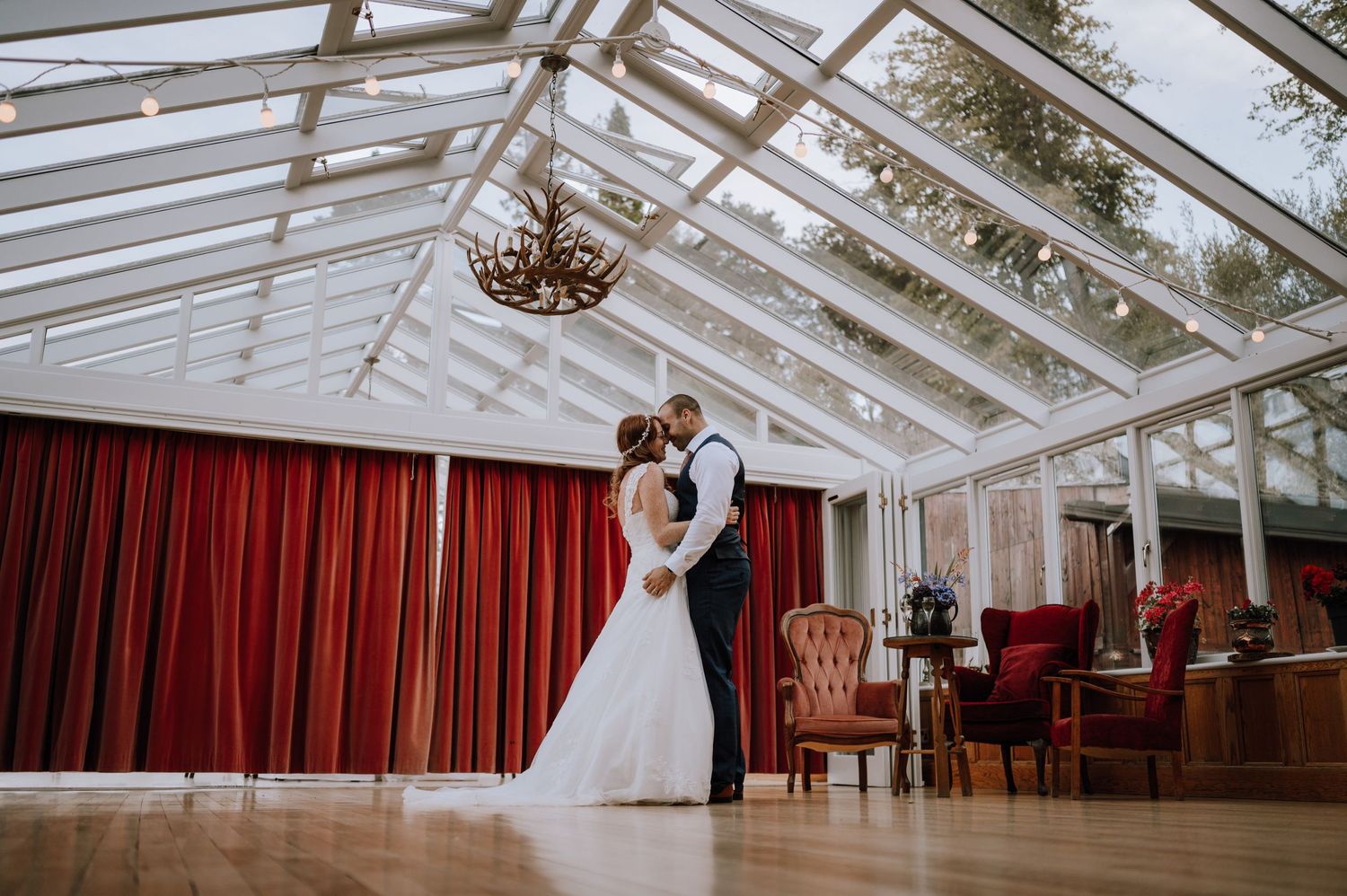 Couple sharing a romantic dance in an elegant glass-roofed wedding venue with red curtains and vintage furniture.