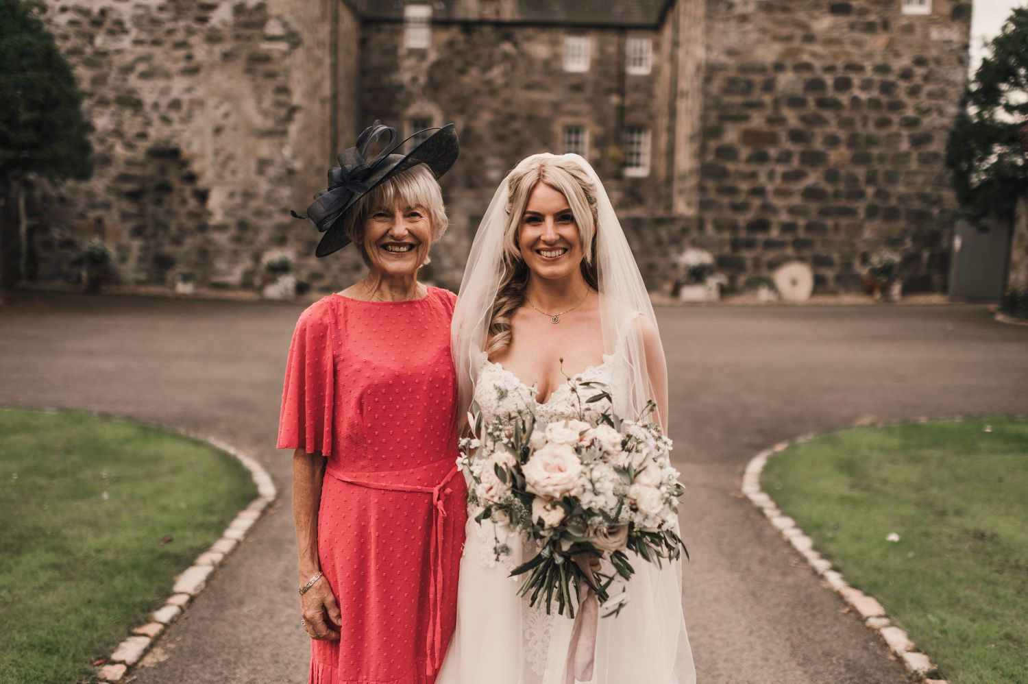 Two wedding guests pose together against a stone castle backdrop while holding an elegant white floral bouquet.