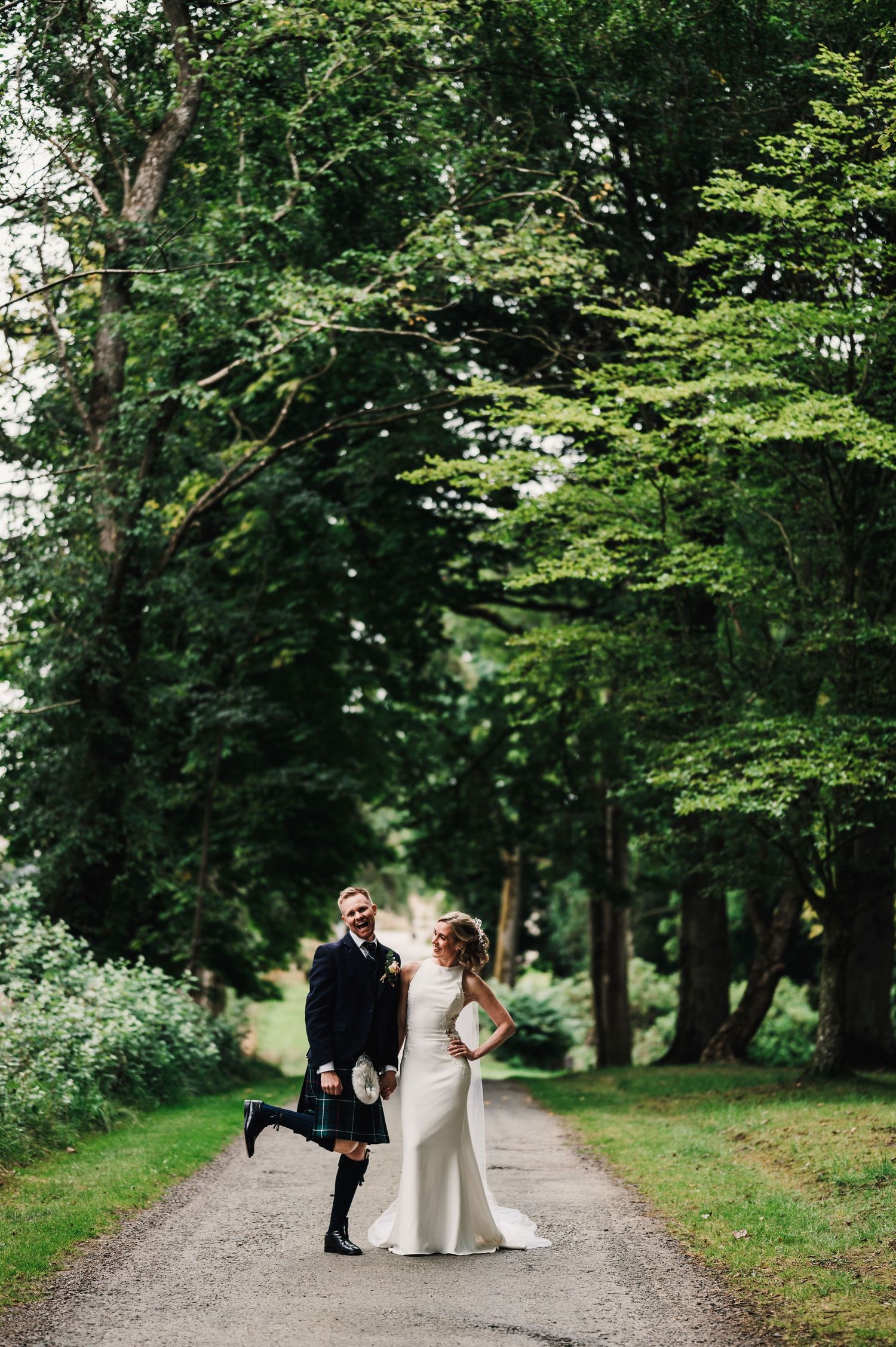 Wedding couple posing playfully on a tree-lined path with the groom wearing traditional Scottish kilt.
