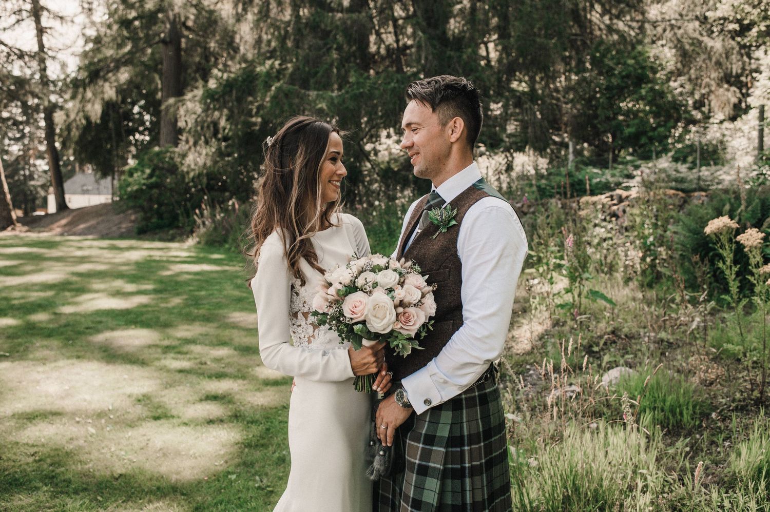 Romantic wedding couple in Scottish attire poses with pink and white bouquet in woodland setting.