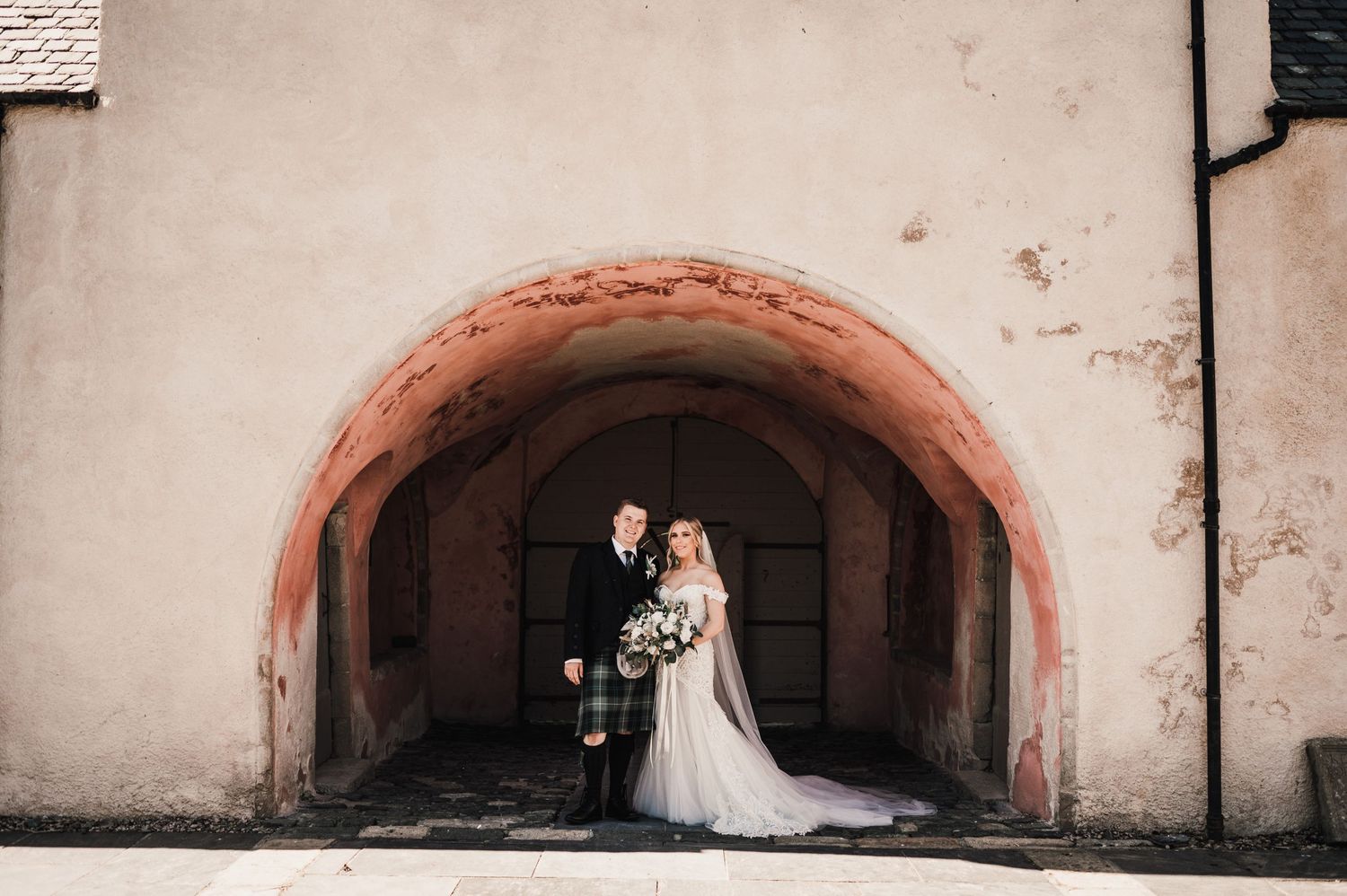 A wedding couple poses under a rustic stone archway creating a dramatic framed portrait.