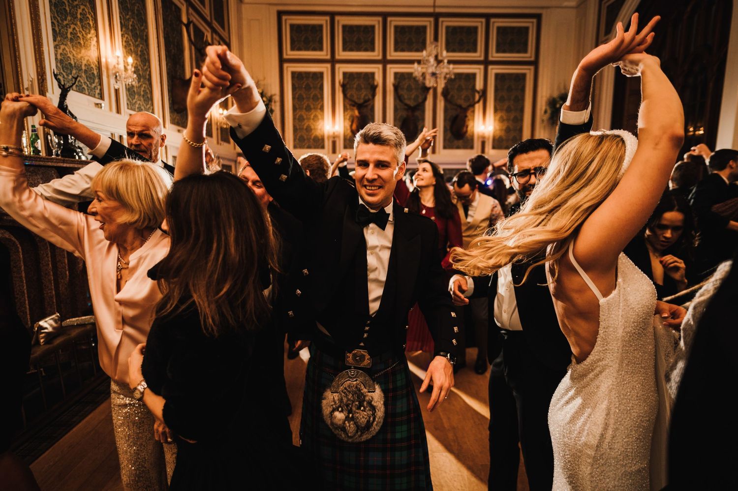 Wedding celebration with guests dancing energetically on a crowded dance floor under ornate lighting.