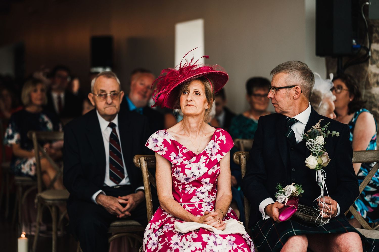 Wedding guests in formal attire seated during ceremony with someone wearing a vibrant pink dress and hat.