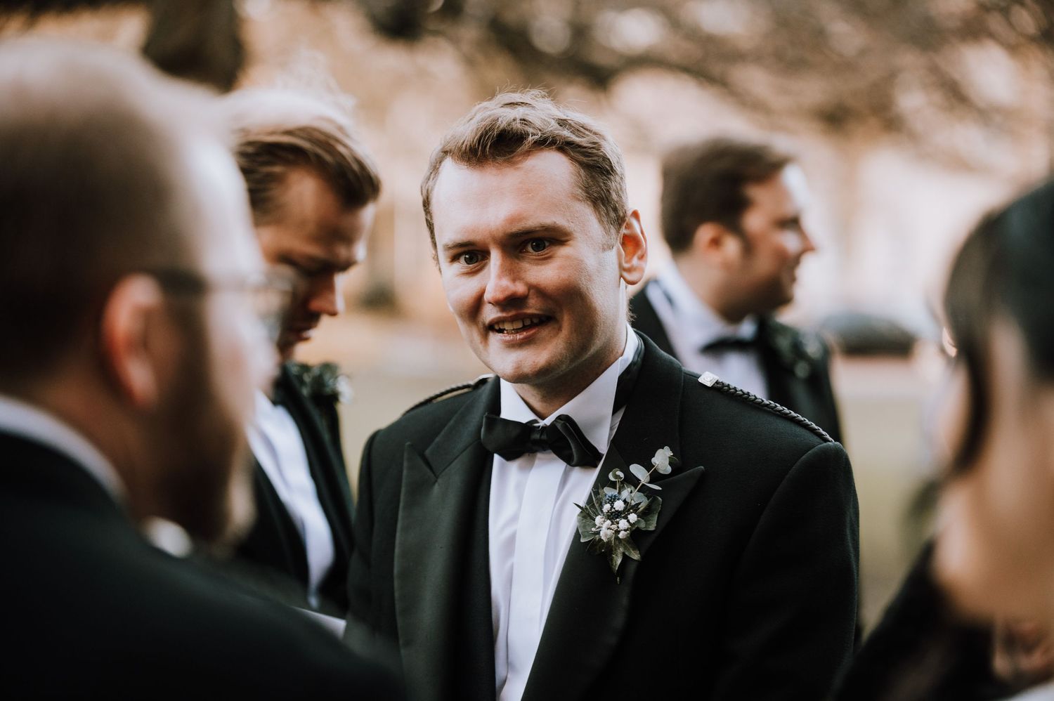 A guest in formal attire smiles during outdoor wedding conversations.