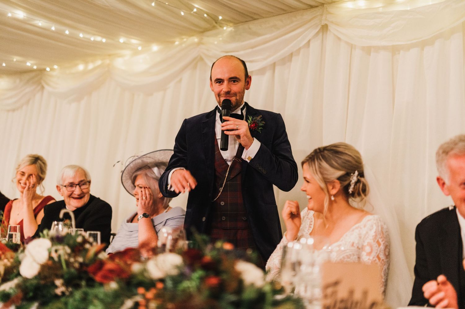 A speaker delivers a speech at an elegant wedding reception with white tent draping and string lights.