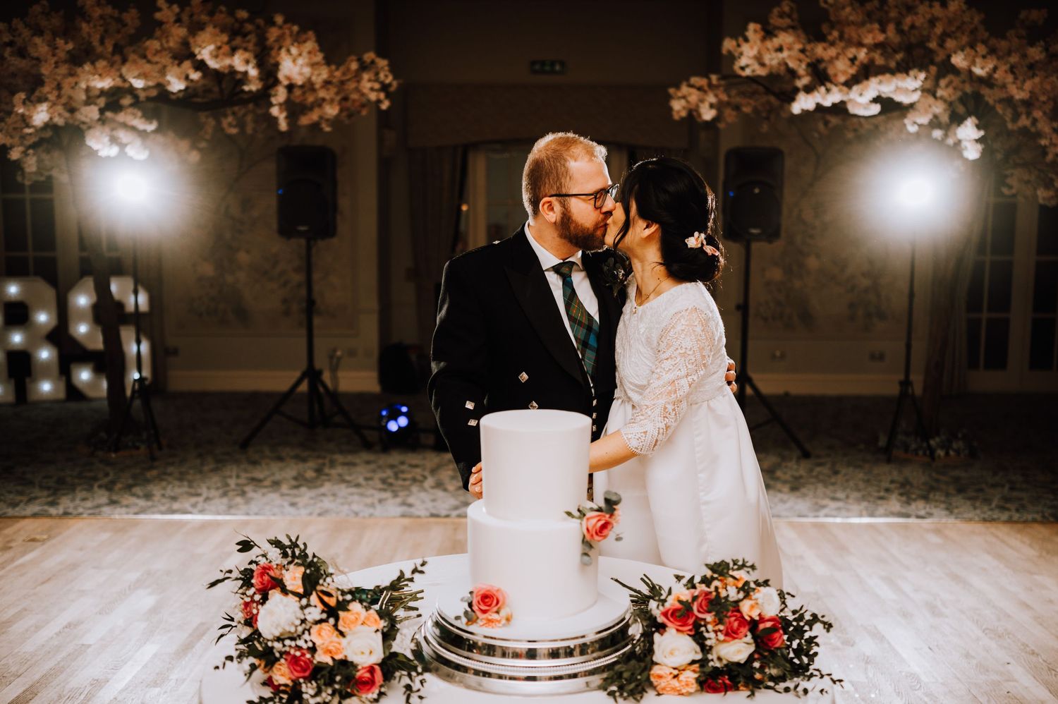 Couple sharing intimate moment while cutting elegant white wedding cake with floral accents.