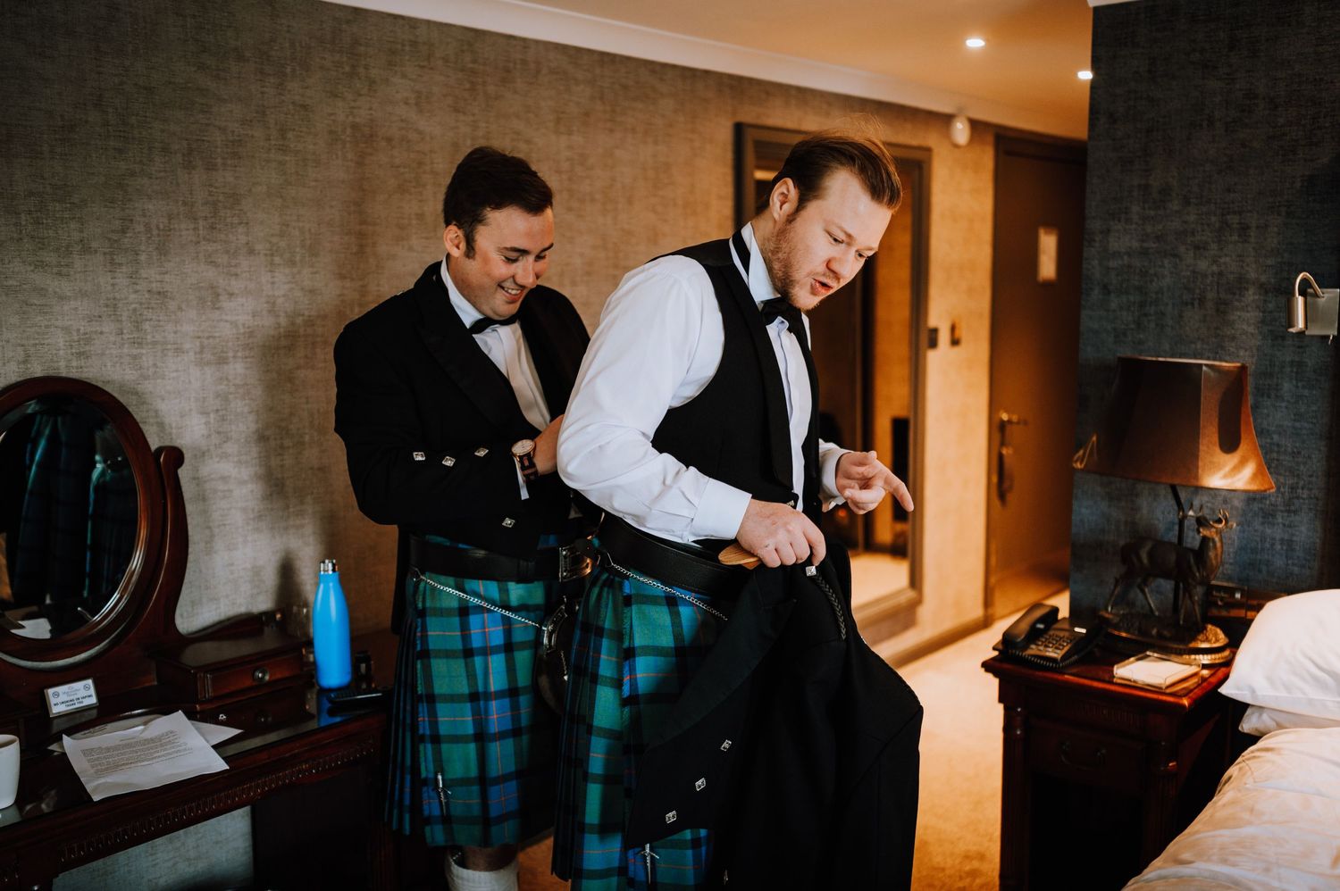 Two people getting dressed in traditional Scottish kilts and formal wear in a hotel room.