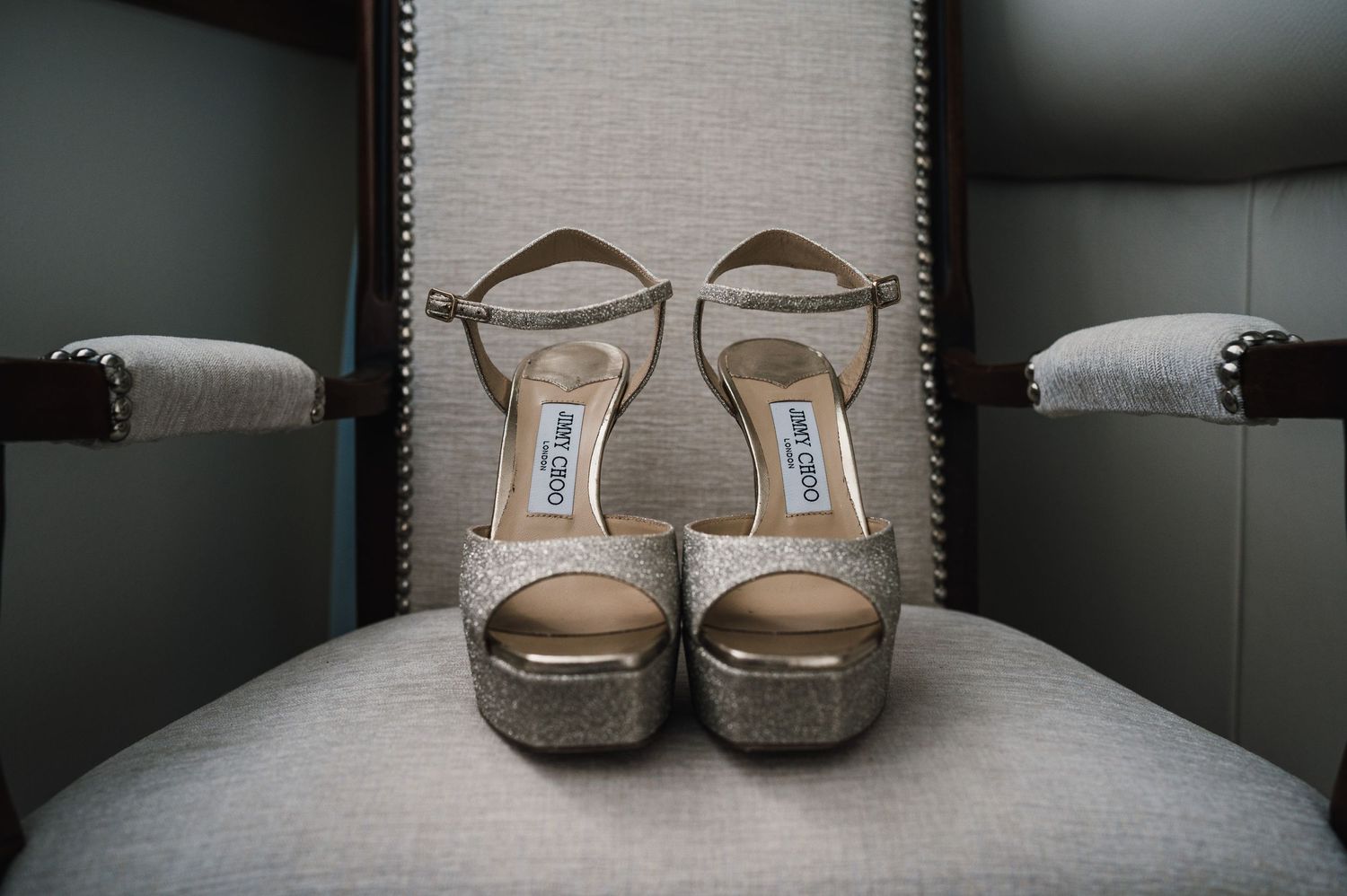 Gold metallic Jimmy Choo platform sandals sitting on a gray upholstered chair.