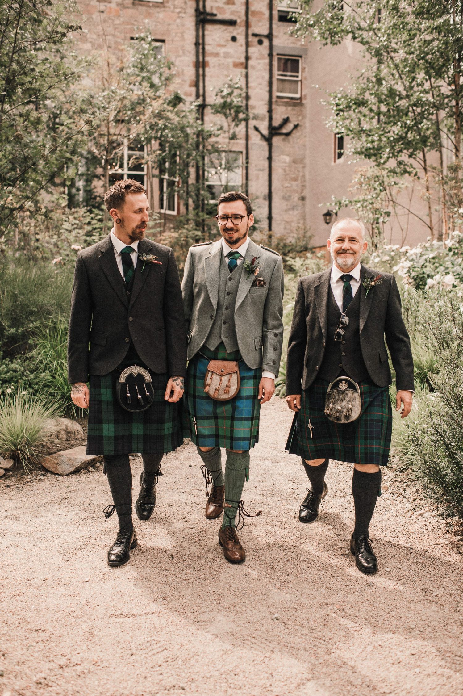 Three people in kilts and formal jackets walking along a garden path by a brick building.